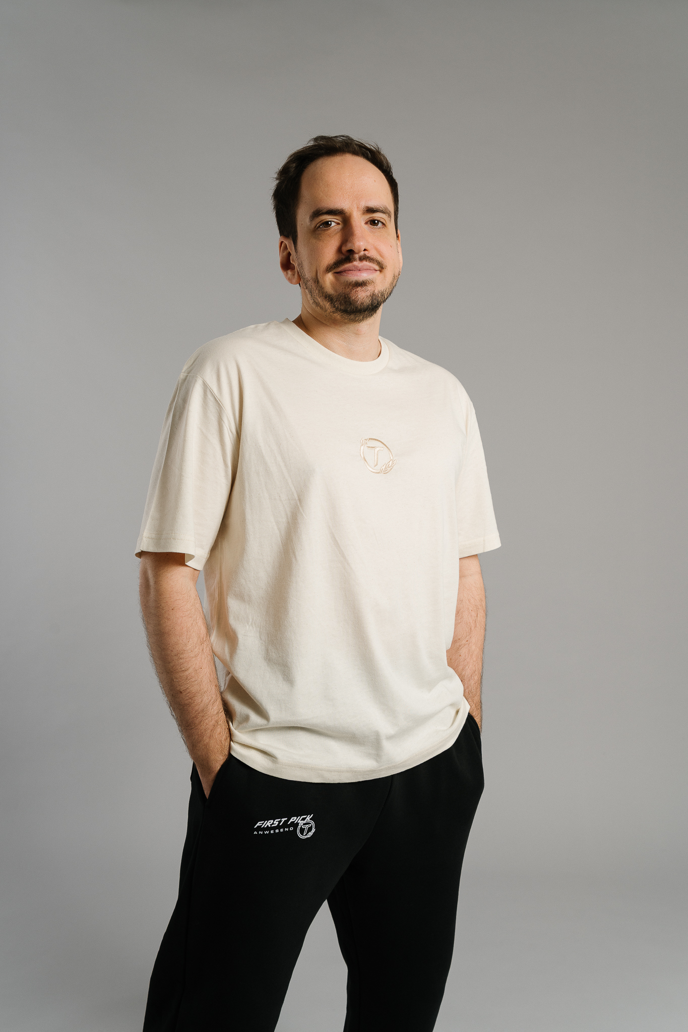 Eine Person trägt ein beiges T-Shirt mit dezentem, hell gesticktem Logo auf der Brust – ein rundes Emblem mit dem Buchstaben „T“. Das Shirt hat einen lockeren Schnitt und kurze Ärmel. Die Person steht locker mit den Händen in den Hosentaschen und lächelt leicht in neutraler Studio-Umgebung.