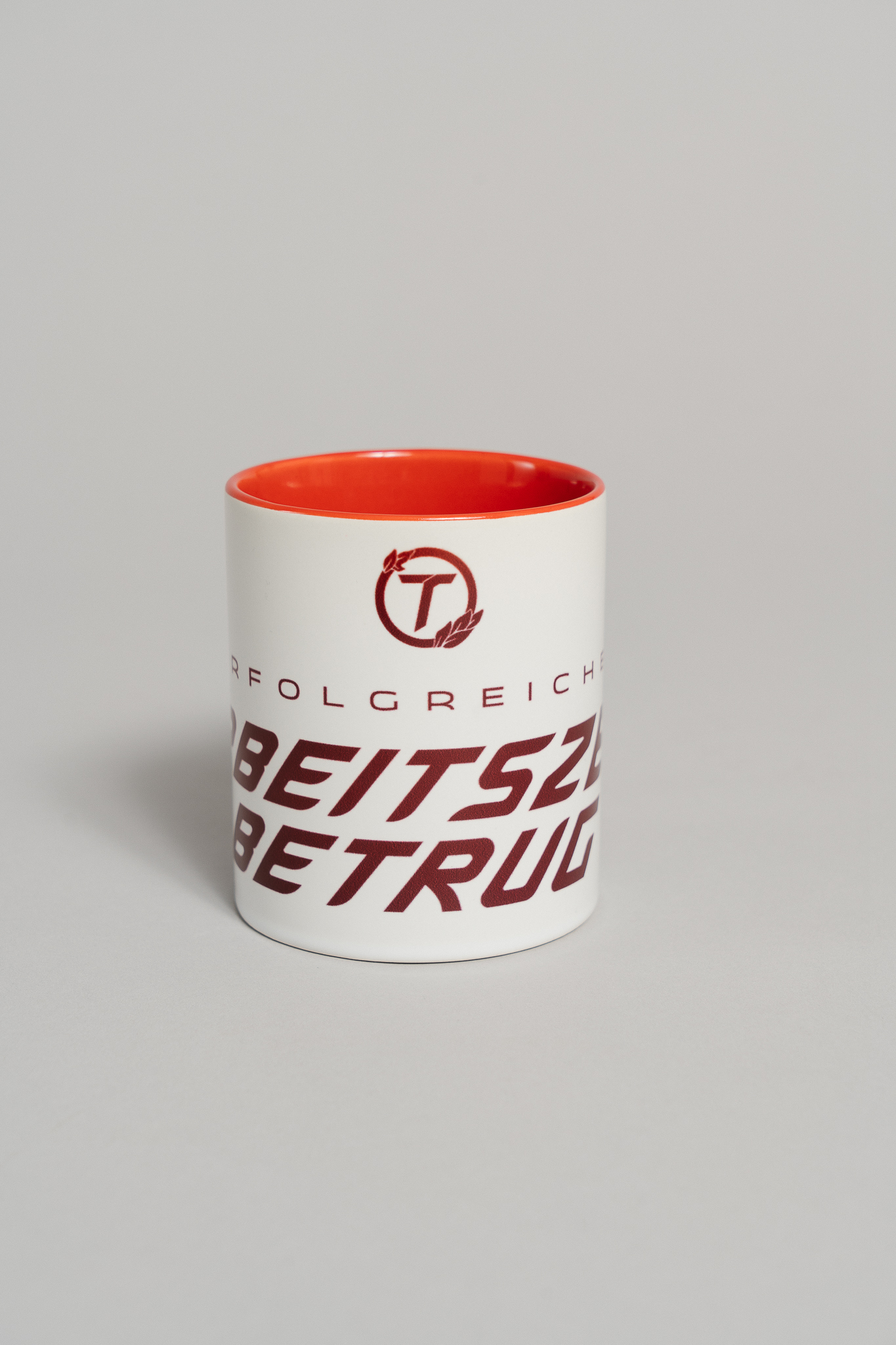 Weiße Keramiktasse mit rotem Innenbereich und rotem Schriftzug. Auf der Vorderseite steht in markanter, schräger Schrift der Text „ERFOLGREICHER ARBEITSZEITBETRUG“. Darüber befindet sich ein rundes Logo mit einem stilisierten Buchstaben „T“ und zwei Blättern. Die Tasse steht auf einer neutral grauen Fläche.
