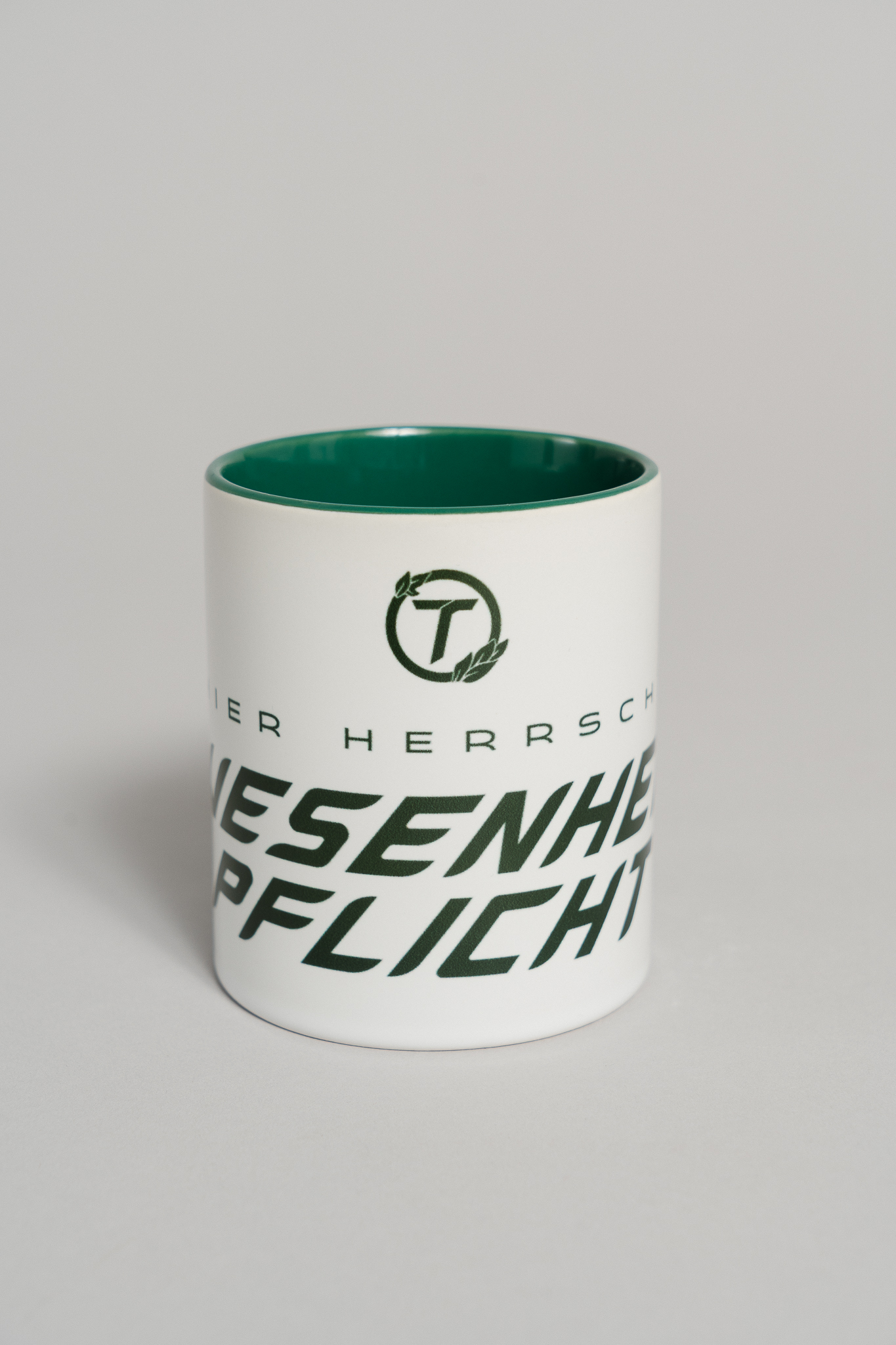 Weiße Keramiktasse mit dunkelgrünem Innenleben. Auf der Vorderseite ist ein großer, schräg gesetzter Schriftzug in dunklem Grün zu sehen, der teilweise den Text „HIER HERRSCHT ANWESENHEITSPFLICHT“ zeigt. Über dem Text befindet sich ein rundes Logo mit dem Buchstaben „T“ und zwei Blättern als feines grafisches Element. Die Tasse steht auf einer neutralen grauen Fläche.