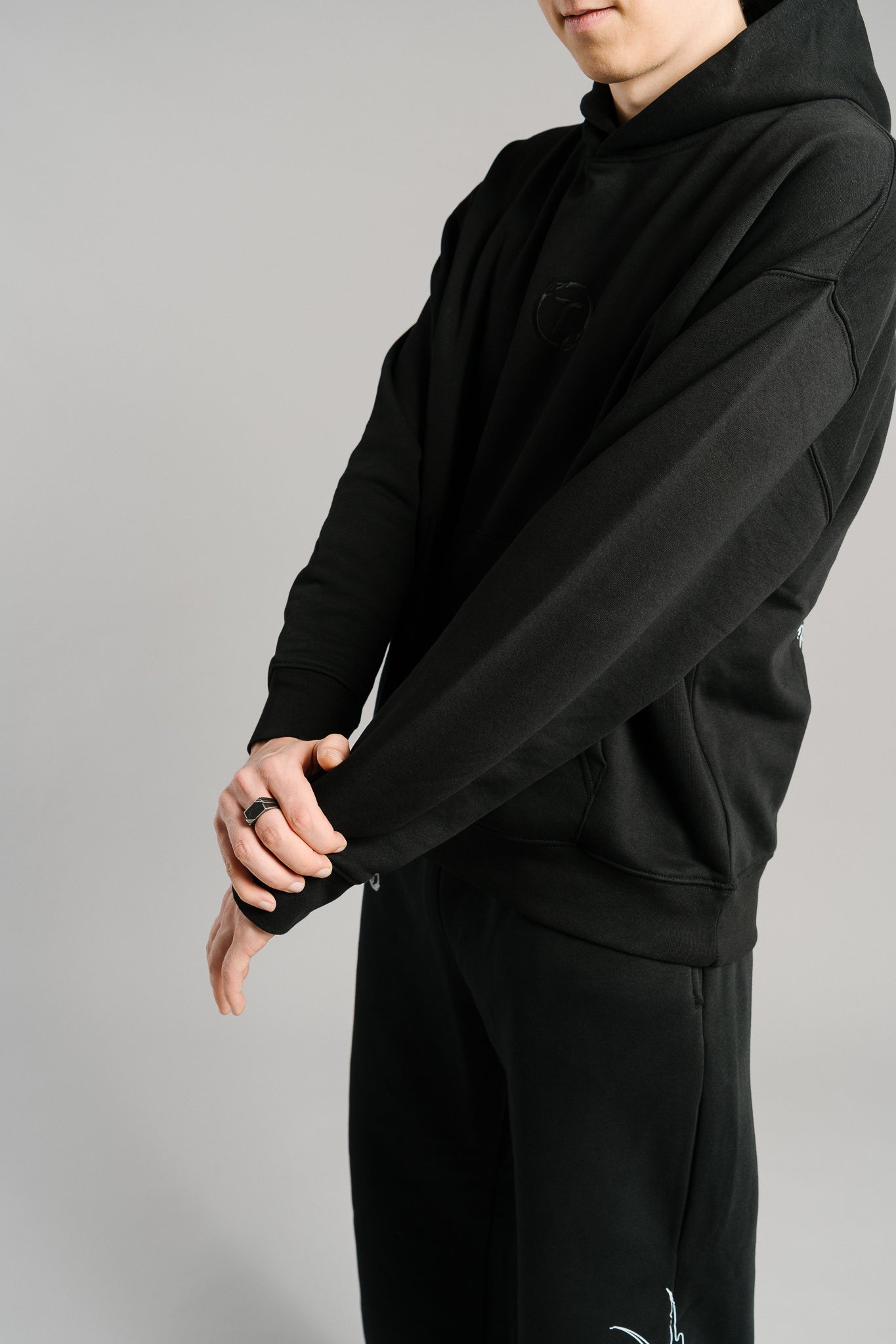 Schwarzer Hoodie aus weichem Stoff mit dezentem Ton-in-Ton-Sticklogo auf der Brust. Die Nahaufnahme zeigt den langen Ärmel und die strukturierte Oberfläche des Stoffes. Das schlichte Design mit Kängurutasche und lockerer Passform vermittelt einen minimalistischen, hochwertigen Streetwear-Look.