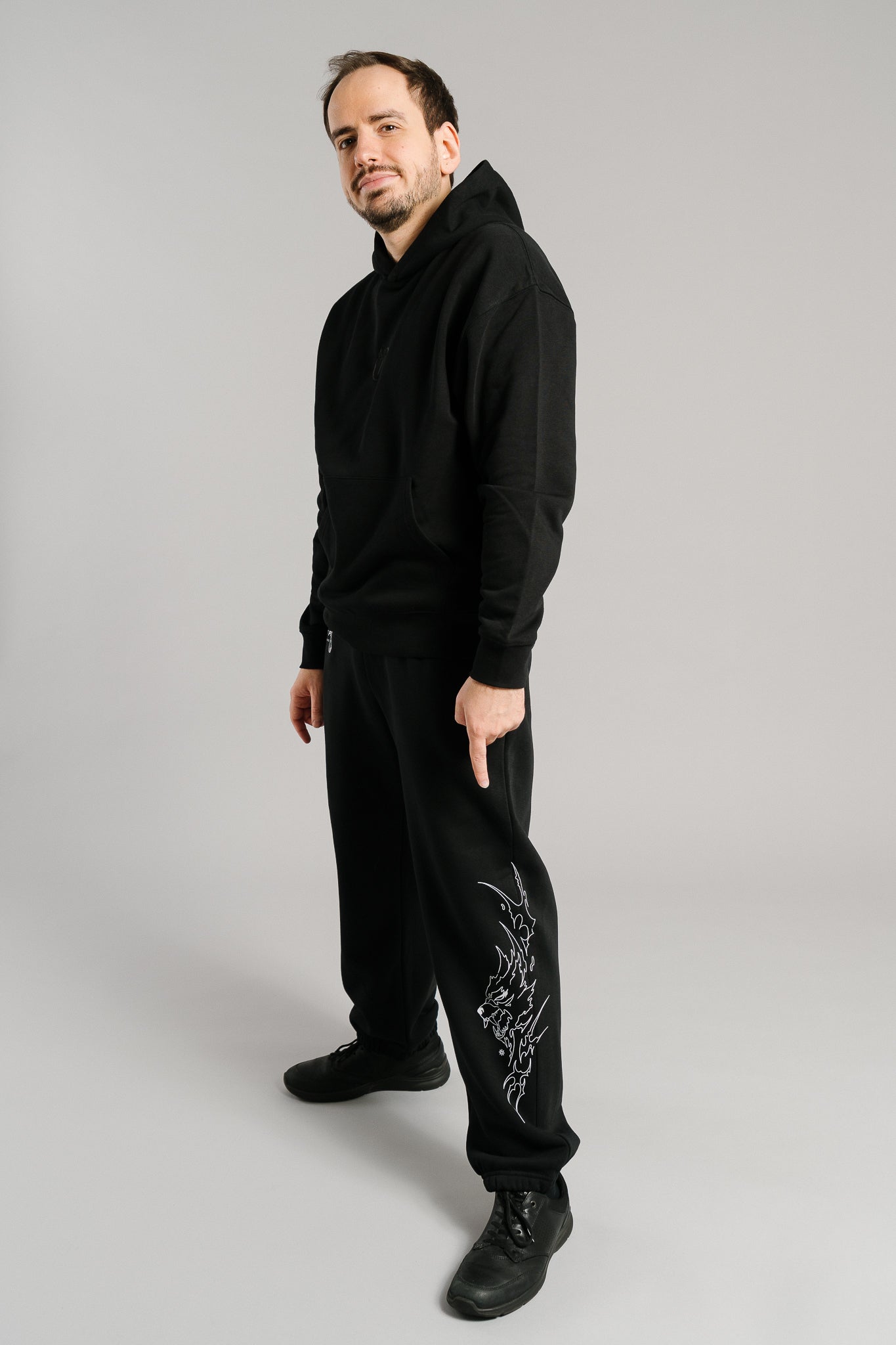 Schwarze Jogginghose mit weißem Flammenmotiv in Form eines stilisierten Tierkopfes auf dem rechten Bein. Der Print verläuft vertikal entlang der äußeren Beinseite. Die Hose hat eine lockere Passform, elastische Bündchen an den Knöcheln und besteht aus weichem Sweatstoff. Auf dem linken Oberschenkel ist zusätzlich der weiße Schriftzug „FIRST PICK ANWESEND“ mit rundem Logo sichtbar.