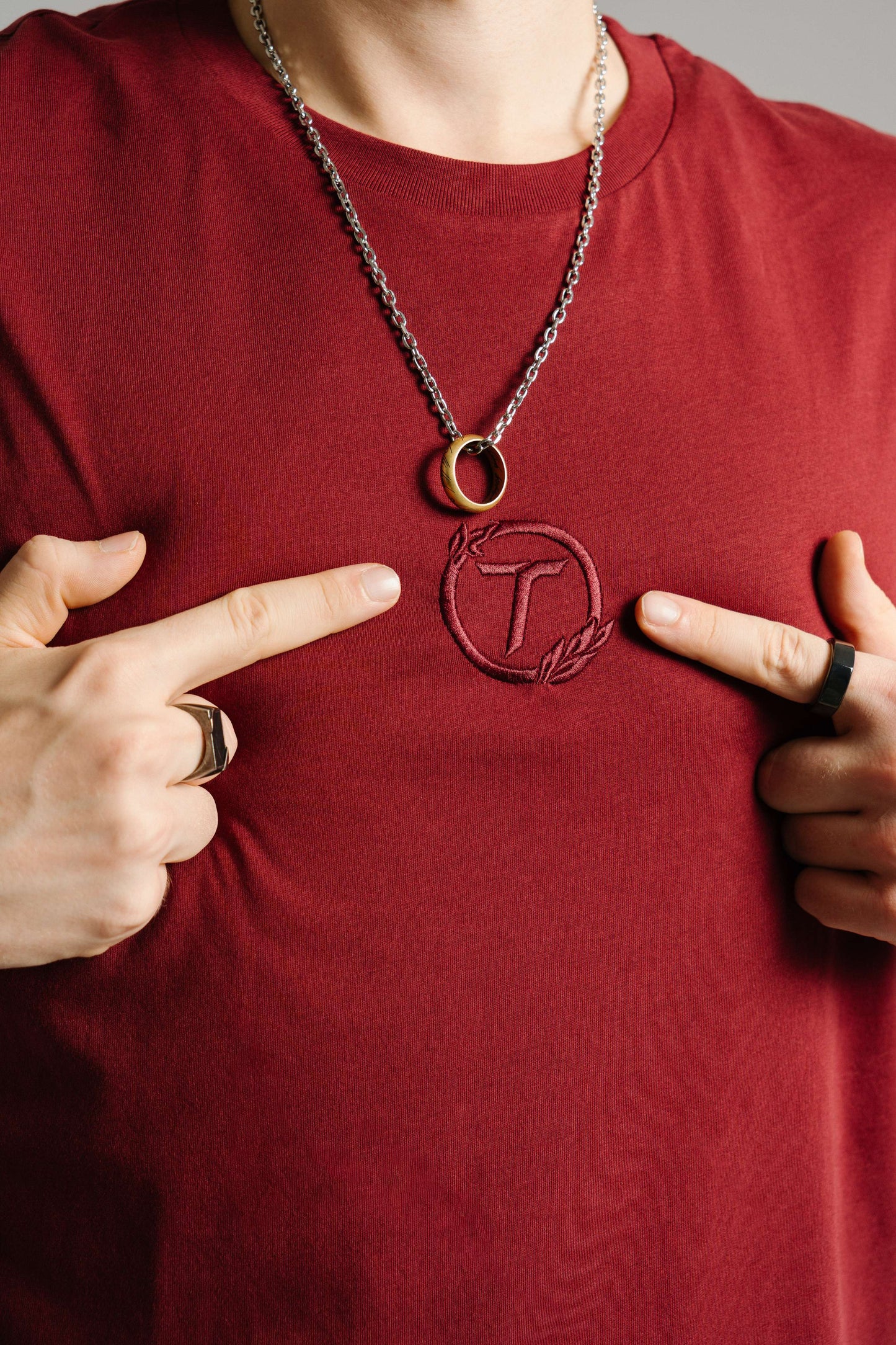 Nahaufnahme eines burgunderroten T-Shirts aus Baumwolle mit tonaler Stickerei des „T“-Logos im Lorbeerkranz auf der Brust. Zwei Finger zeigen auf das gestickte Emblem, das mittig unterhalb einer silbernen Halskette mit goldenem Ringanhänger liegt. Der Fokus liegt auf der Logo-Stickerei und der feinen Stoffstruktur.