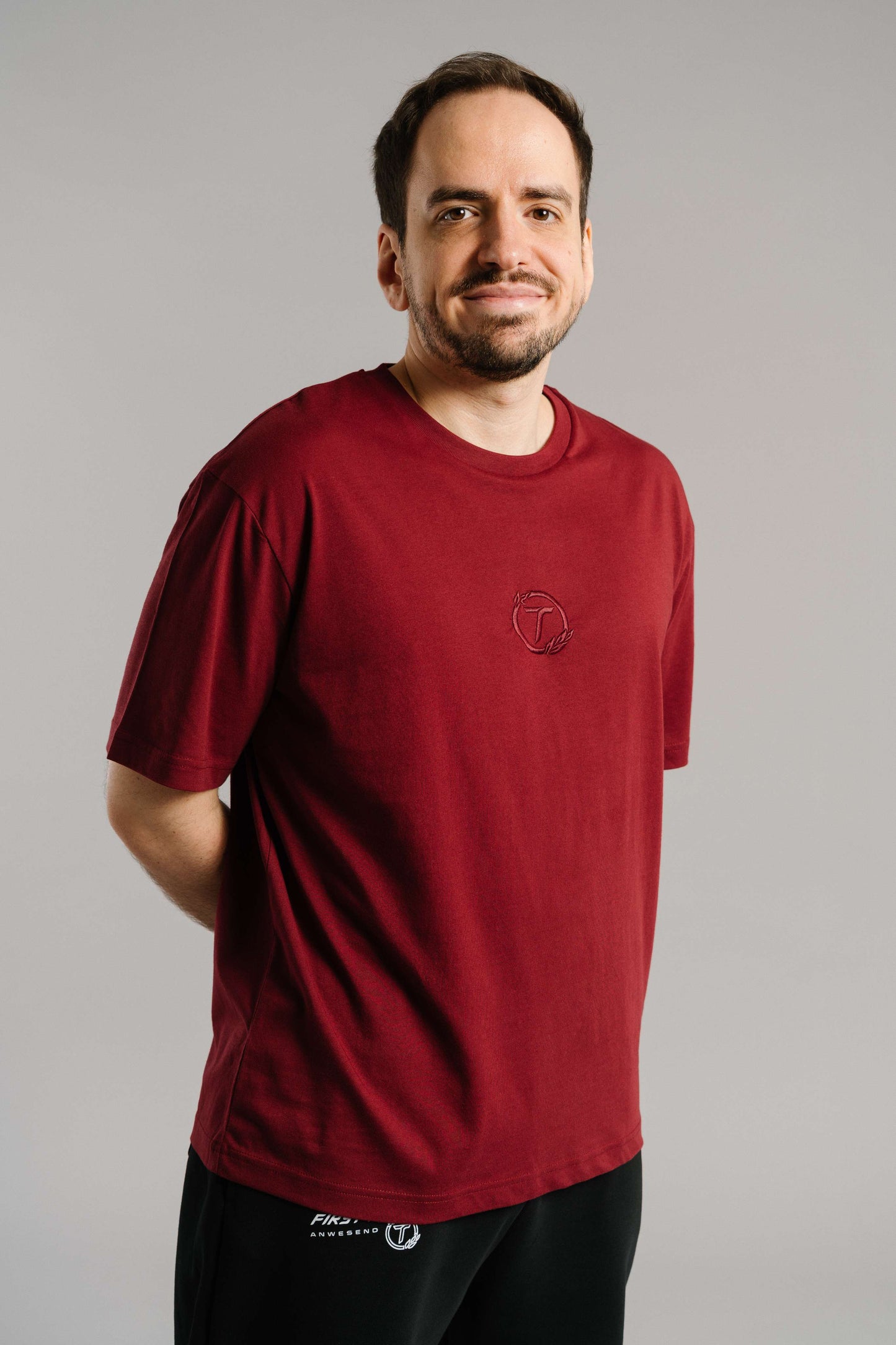 Ein burgunderrotes T-Shirt mit tonaler Stickerei des „T“-Logos im Lorbeerkranz auf der Brust. Das Shirt hat einen geraden Schnitt, kurze Ärmel und besteht aus weicher Baumwolle. Es wird locker getragen und vermittelt einen cleanen, minimalistischen Look.