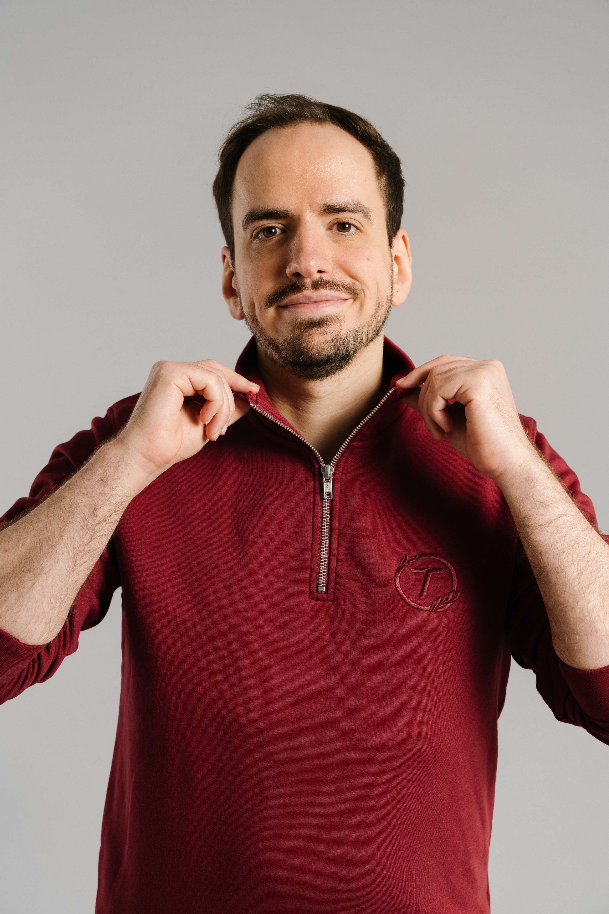 Ein burgunderrotes Sweatshirt mit kurzem Reißverschluss am Kragen. Auf der linken Brust ist das ton-in-ton gestickte Logo mit dem Buchstaben „T“ im Lorbeerkranz zu sehen. Der Stoff wirkt weich und hochwertig. Die Person trägt das Sweatshirt offen am Kragen und hält mit beiden Händen den Kragen leicht nach außen, während sie freundlich in die Kamera blickt.