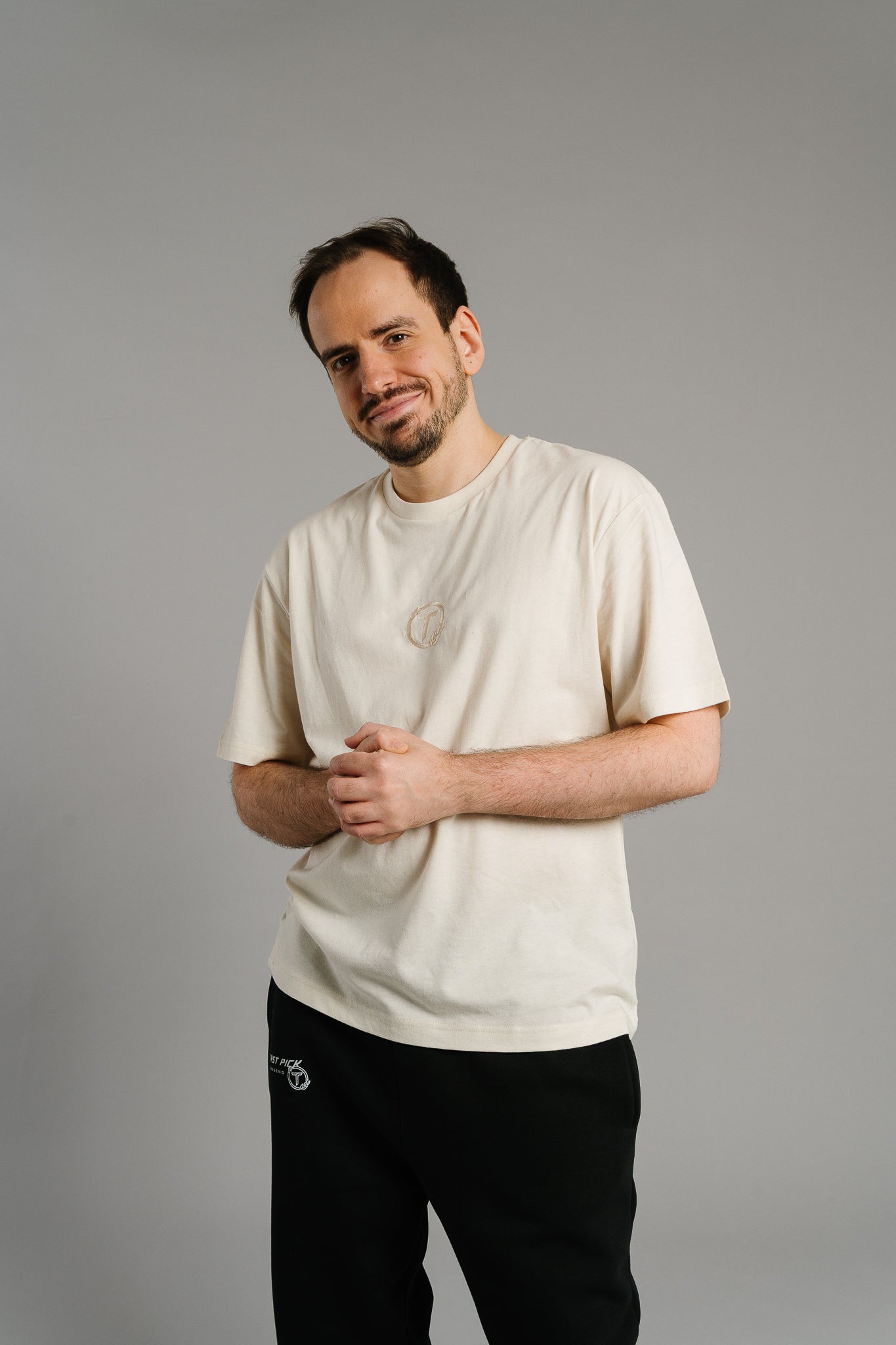 Eine Person trägt ein beiges T-Shirt mit kleinem, dezent gesticktem Logo auf der Brust. Das Logo zeigt den Buchstaben „T“ in einem Kreis mit Lorbeerblättern. Das weiche Material und der entspannte Schnitt verleihen dem Shirt eine lässige, hochwertige Ausstrahlung. Die Person steht locker mit gefalteten Händen und lächelt leicht in die Kamera.