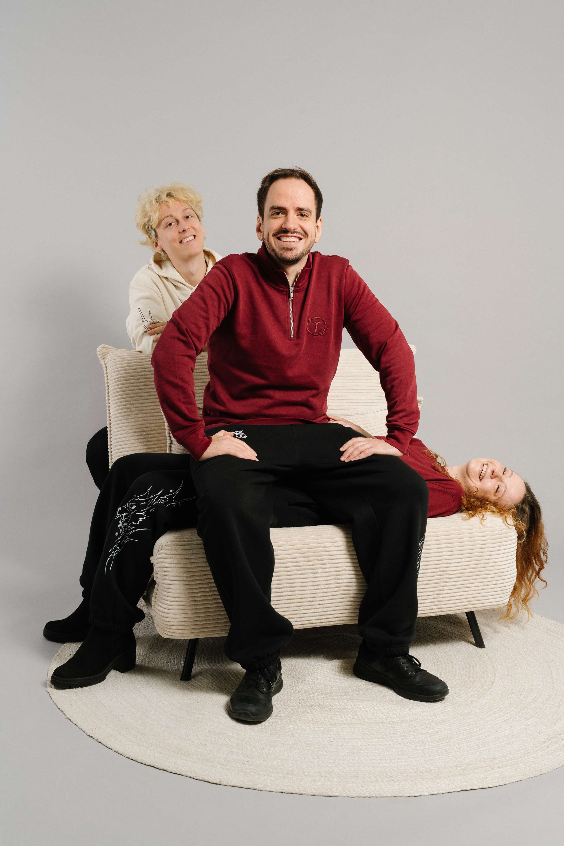Ein burgunderrotes Sweatshirt mit kurzem Reißverschluss und gesticktem „T“-Logo auf der Brust. Das Sweatshirt hat eine weiche, strukturierte Oberfläche und wird locker getragen. Die Person sitzt auf einem hellen Sessel, lächelnd und leicht nach vorn gebeugt. Im Hintergrund sitzen zwei weitere Personen, eine in einem cremefarbenen Hoodie und eine in einem gleichfarbigen Sweatshirt, was dem Bild eine dynamische, freundliche Atmosphäre verleiht. Der Fokus liegt auf dem roten Sweatshirt im Vordergrund.