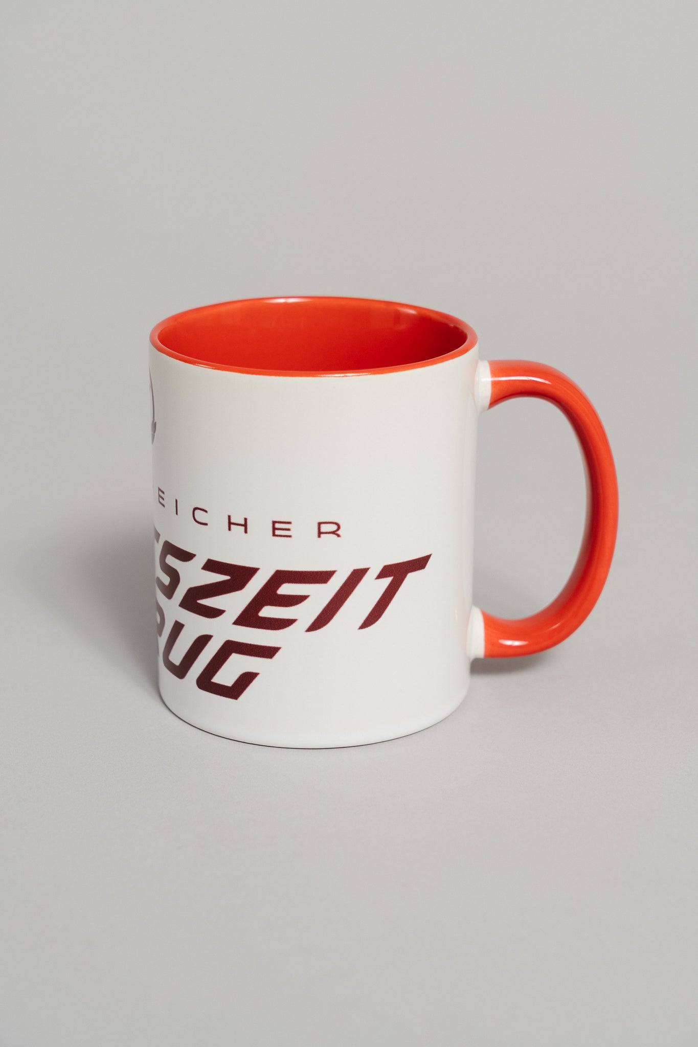 Weiße Keramiktasse mit rotem Henkel und rotem Innenbereich. Auf der Außenseite ist in dunklem Rot ein schräger Schriftzug zu sehen, der teilweise den Text „ERFOLGREICHER ARBEITSZEITBETRUG“ zeigt. Die Tasse steht auf einer neutral grauen Fläche und ist leicht seitlich nach rechts gedreht.