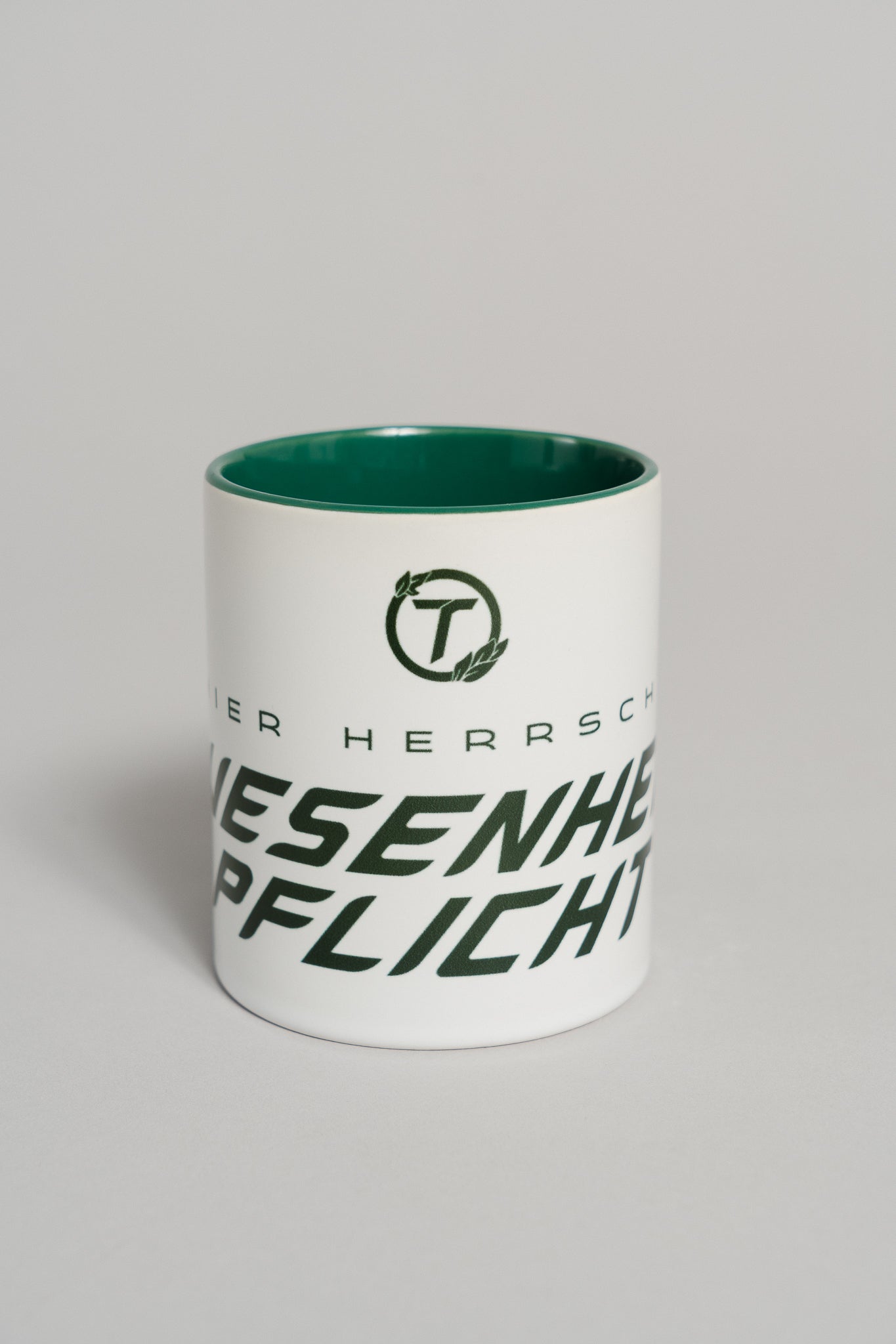 Weiße Keramiktasse mit dunkelgrünem Innenbereich. Auf der Vorderseite ist ein großer, schräg verlaufender Schriftzug in dunklem Grün zu sehen, der teilweise den Text „HIER HERRSCHT ANWESENHEITSPFLICHT“ zeigt. Darüber befindet sich ein rundes Logo mit dem Buchstaben „T“ und kleinen Blattelementen. Die Tasse steht auf einer neutralen grauen Fläche und wird frontal gezeigt.