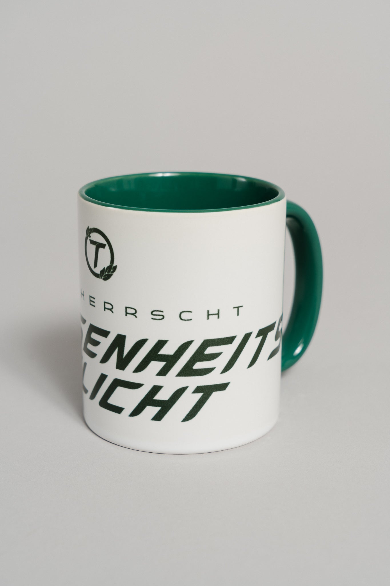 Weiße Keramiktasse mit grünem Henkel und grünem Innenbereich. Auf der Außenseite ist ein dunkler Schriftzug in moderner, schräger Typografie zu sehen, der teilweise den Text „HIER HERRSCHT ANWESENHEITSPFLICHT“ zeigt. Darüber befindet sich ein rundes Logo mit dem Buchstaben „T“ und zwei kleinen Blättern. Die Tasse steht auf einer neutralen, hellgrauen Fläche.