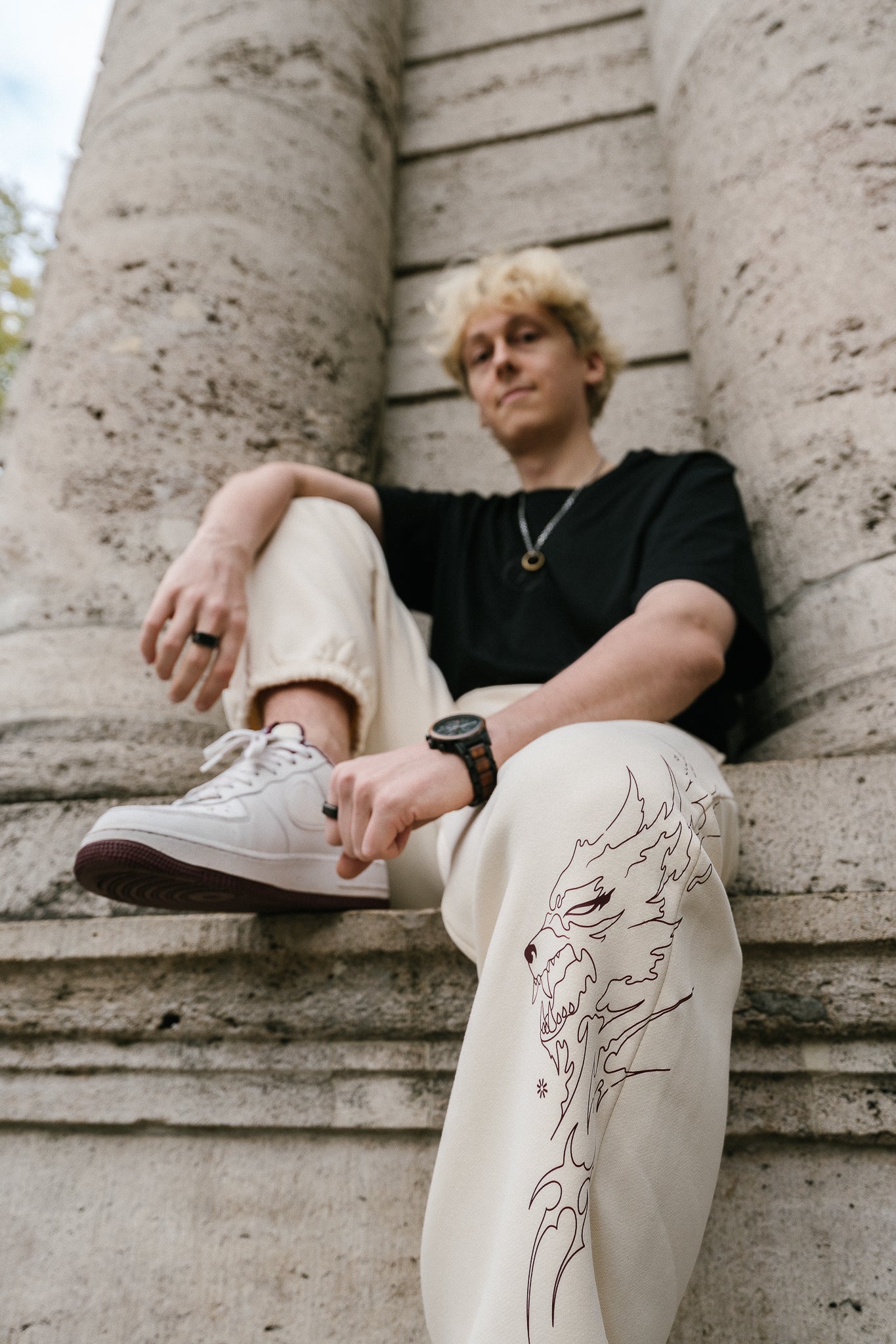 Beigefarbene Jogginghose aus Sweatstoff mit auffälligem, braunrotem Printmotiv auf dem rechten Bein. Der Druck zeigt ein stilisiertes, flammenförmiges Wolfsgesicht in dynamischer Linienführung. Die Hose hat elastische Bündchen an den Knöcheln und wird im Sitzen getragen, wodurch das Motiv gut sichtbar ist.