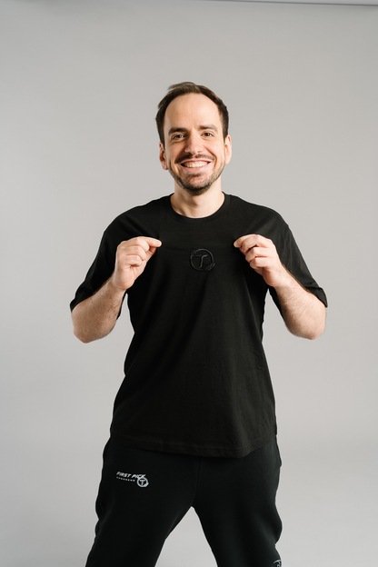 Schwarzes T-Shirt aus weicher Baumwolle mit dezentem Ton-in-Ton-Sticklogo auf der Brust. Das Logo zeigt ein stilisiertes „T“ in einem Kreis mit Lorbeerzweigen. Das Shirt hat einen geraden Schnitt und wirkt schlicht, modern und sportlich – ideal für einen cleanen Streetwear-Look.