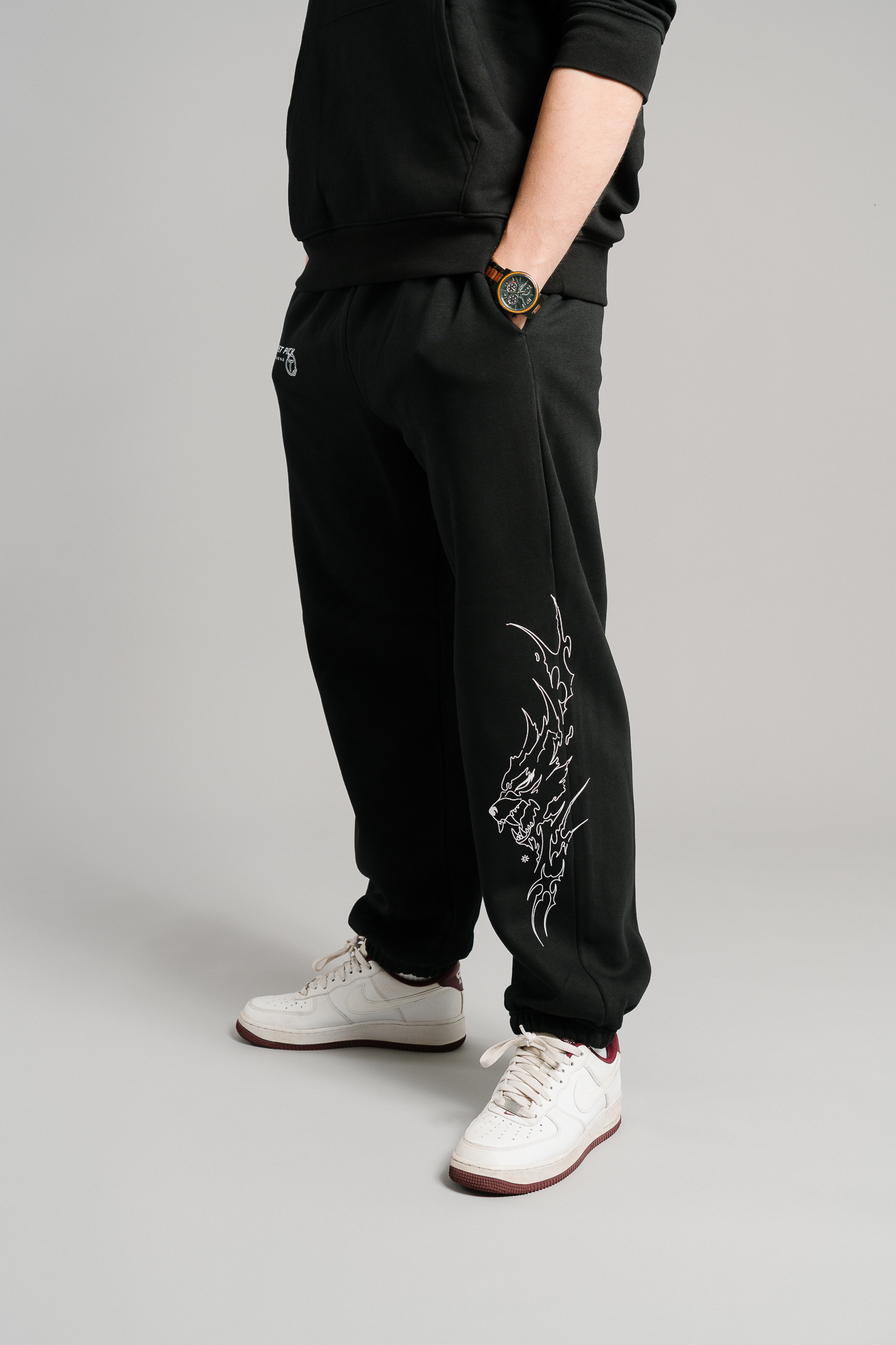 Schwarze Jogginghose mit weißem, grafischem Flammenmotiv entlang des rechten Beins. Das Design zeigt eine stilisierte Flammenlinie mit angedeutetem Tiergesicht. Die Hose verfügt über einen elastischen Bund, seitliche Eingrifftaschen und eine kleine weiße Stickerei mit dem Schriftzug „FIRST PICK ANWESEND“ samt Logo oberhalb des linken Oberschenkels. Locker geschnitten aus weichem Sweatstoff.