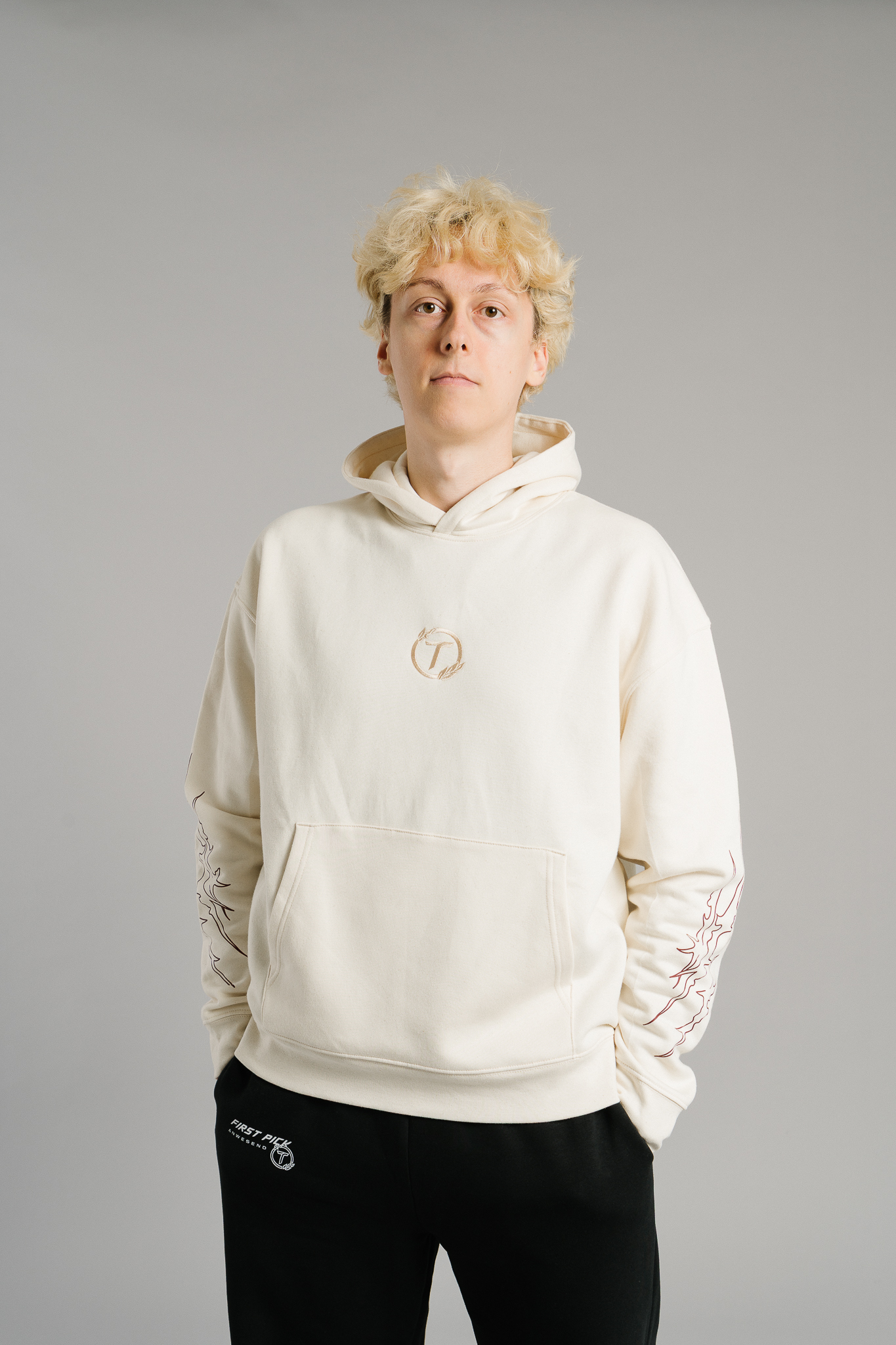 Beigefarbener Hoodie aus Sweatstoff mit Kängurutasche und lockerem Schnitt. Auf der Brust befindet sich ein kleines rundes Logo in Braun mit stilisiertem Buchstaben „T“. Beide Ärmel zeigen braunrote, flammenartige Linienmotive entlang der Außenseiten.