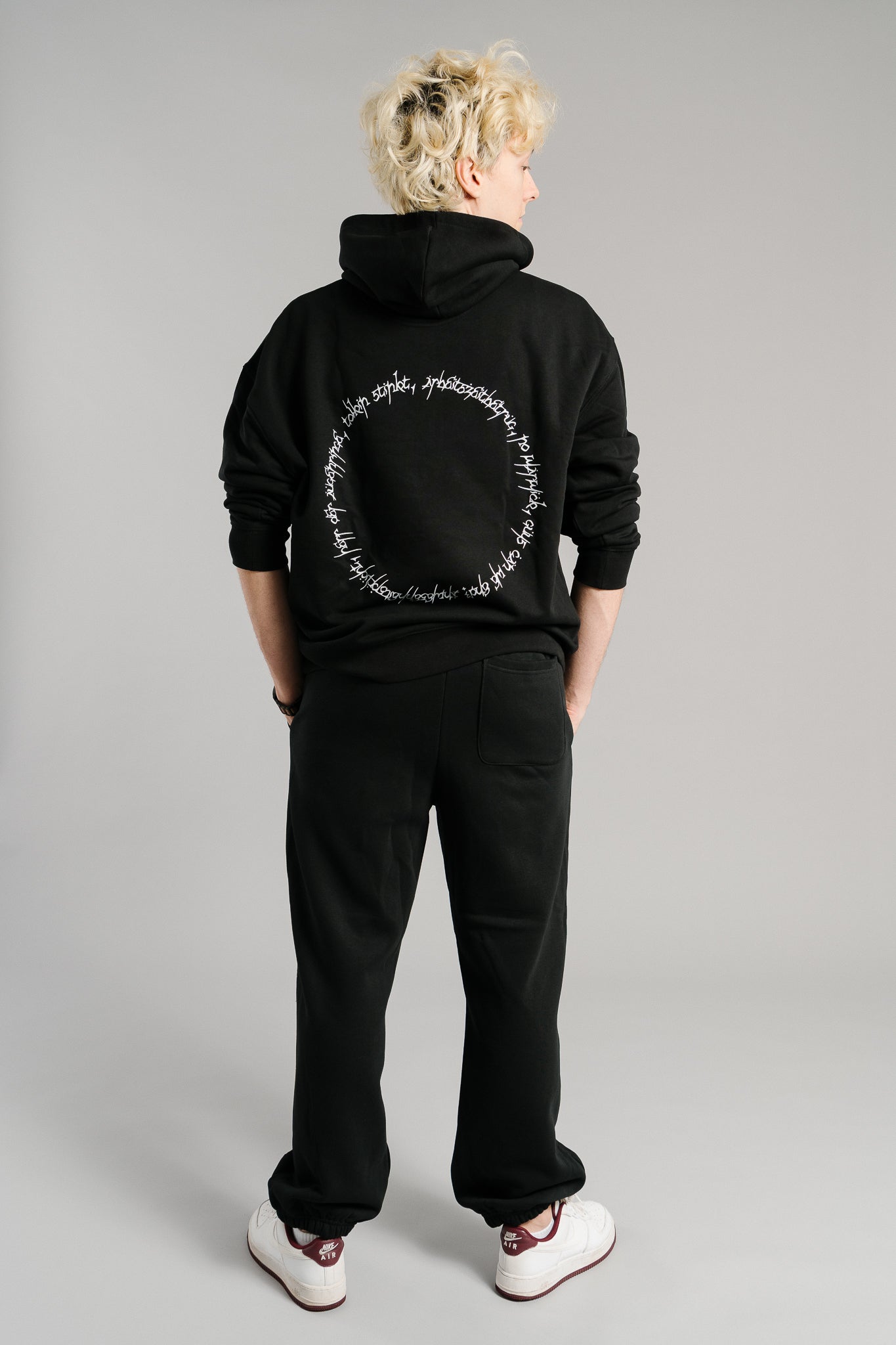 Schwarzer Hoodie mit großem weißen Schriftzug-Print auf der Rückenpartie. Das Design zeigt einen kreisförmig angeordneten Text in handschriftlicher Typografie. Der Kapuzenpullover besteht aus weichem Material, hat eine lockere Passform und betont durch das schlichte Schwarz-Weiß-Kontrastdesign einen modernen Streetwear-Look.
