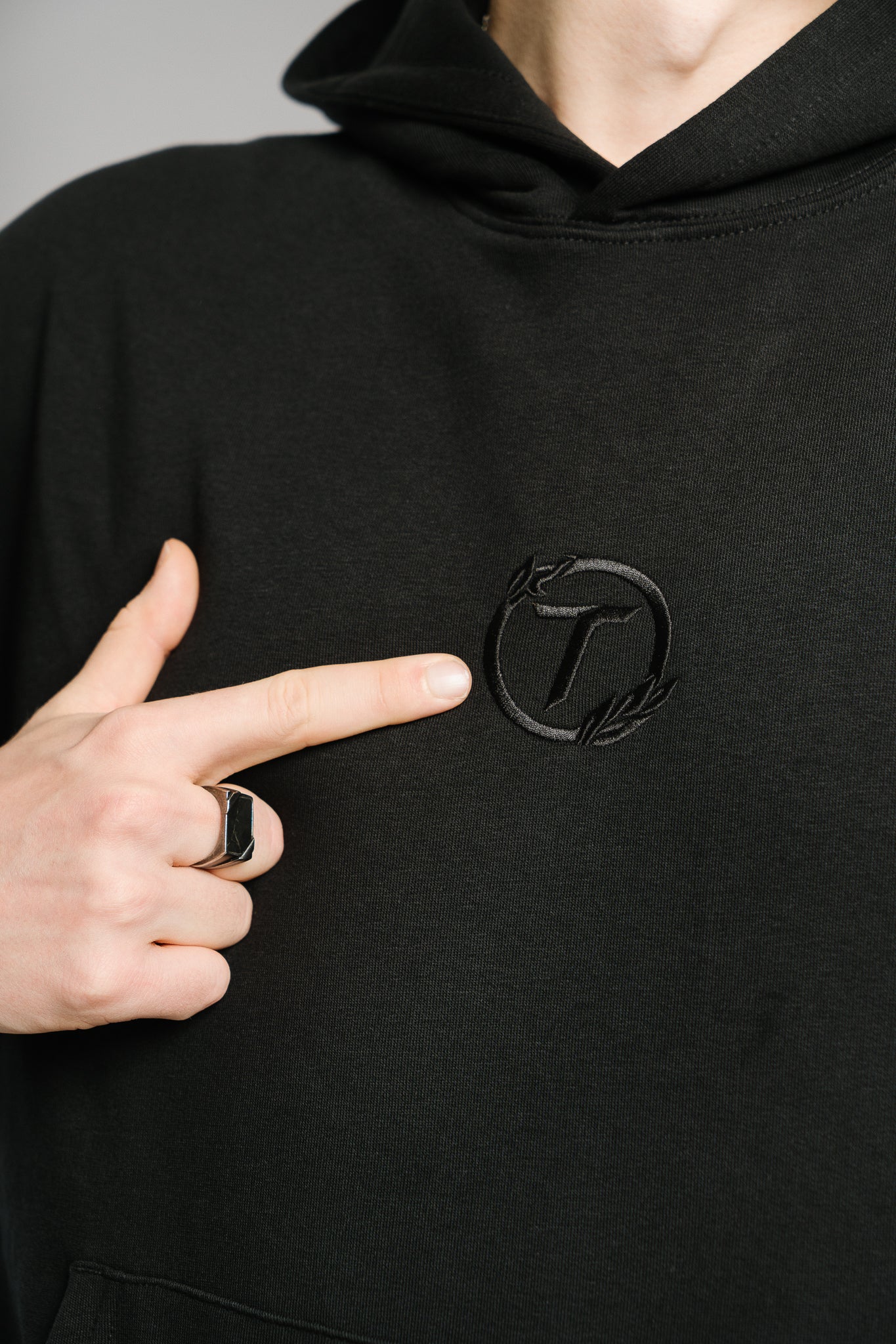 Nahaufnahme eines schwarzen Hoodies mit Ton-in-Ton-Stickerei auf der Brust. Die Stickerei zeigt ein stilisiertes „T“ in einem runden Lorbeerkranz. Der hochwertige, matte Stoff unterstreicht den minimalistischen Look des Kapuzenpullovers.
