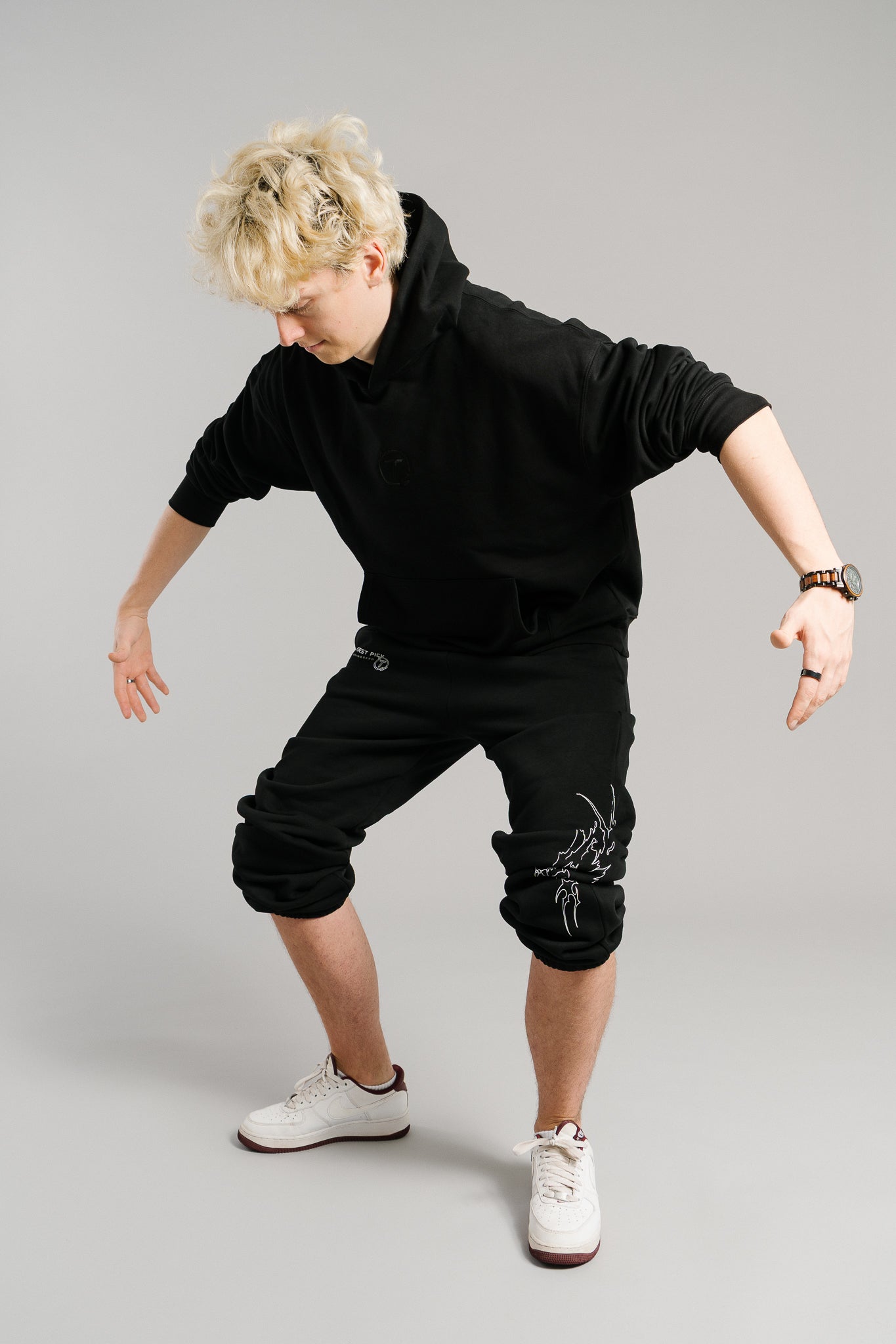 Schwarze Jogginghose mit hochgezogenen Hosenbeinen, wodurch die weißen Sneaker sichtbar sind. Auf der linken Seite befindet sich oberhalb des Knies ein weißer Schriftzug „FIRST PICK ANWESEND“ mit kleinem Rundlogo. Weiter unten am Bein ist ein großflächiger, weißer Flammen-Print in Form eines stilisierten Tierkopfes zu sehen. Die Hose besteht aus weichem Sweatstoff mit elastischem Bund und sportlicher Passform.