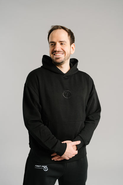 Schwarzer Hoodie aus weichem Baumwollstoff mit dezenter Ton-in-Ton-Stickerei eines stilisierten „T“ im Lorbeerkranz auf der Brust. Das Design ist schlicht, modern und hochwertig verarbeitet, mit klassischer Kängurutasche und Kapuze. Der Pullover steht für einen cleanen Streetwear-Look mit sportlicher Note.