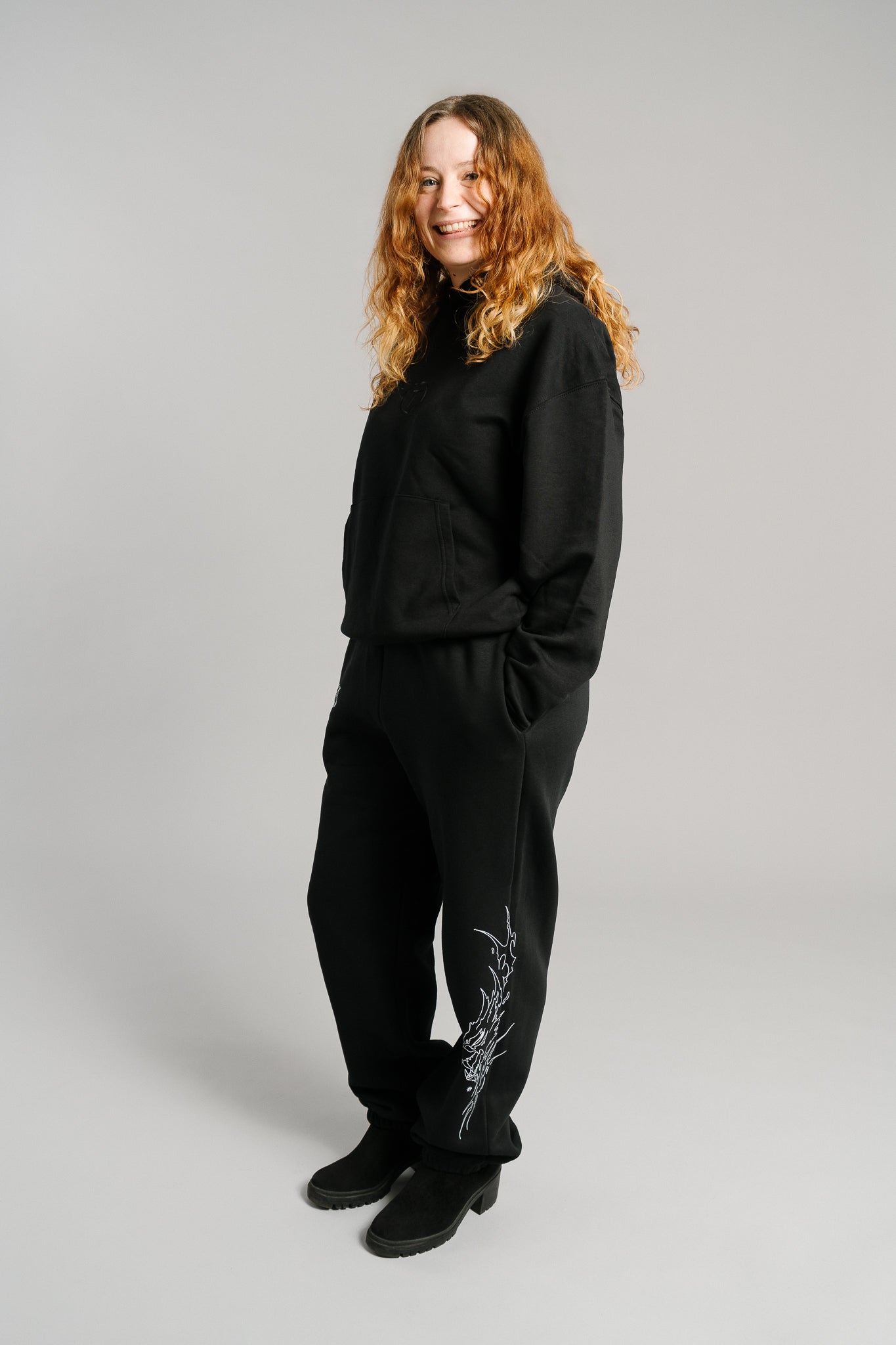 Schwarze Jogginghose mit weißem Flammenmotiv an der Außenseite des rechten Beins. Das Design zeigt eine stilisierte, dynamisch gezeichnete Tierkopf-Illustration in Linienoptik. Die Hose hat eine lockere Passform mit elastischen Bündchen an den Knöcheln und besteht aus weichem, blickdichtem Sweatstoff.