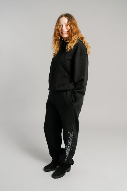 Schwarze Jogginghose mit weißem Flammenmotiv an der Außenseite des rechten Beins. Das Design zeigt eine stilisierte, dynamisch gezeichnete Tierkopf-Illustration in Linienoptik. Die Hose hat eine lockere Passform mit elastischen Bündchen an den Knöcheln und besteht aus weichem, blickdichtem Sweatstoff.