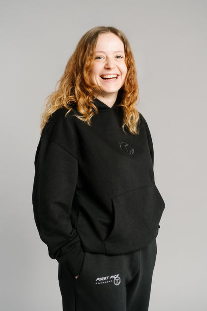 Schwarzer Hoodie mit minimalistischer Ton-in-Ton-Stickerei eines stilisierten „T“ im Lorbeerkranz auf der Brust. Der Pullover besteht aus weichem, leicht strukturiertem Stoff, hat eine große Kängurutasche und eine Kapuze mit lockerer Passform. Das Design wirkt schlicht, modern und unisex.