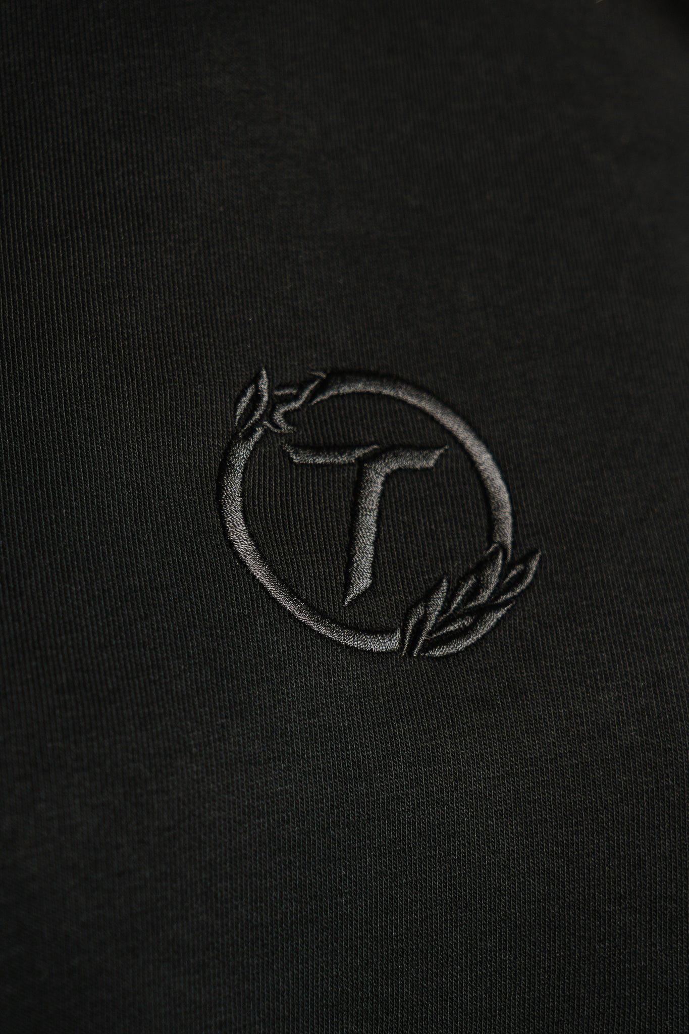 Detailaufnahme eines schwarzen Hoodies mit feiner Ton-in-Ton-Stickerei. Das gestickte Emblem zeigt ein stilisiertes „T“ in einem kreisförmigen Lorbeerkranz. Die Nahaufnahme hebt die strukturierte Stoffoberfläche und die hochwertige Stickqualität hervor.