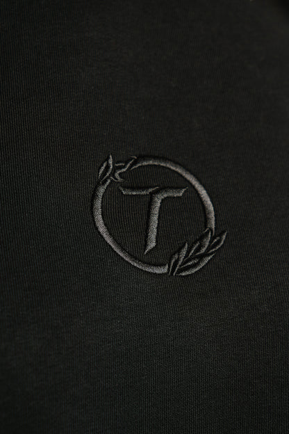 Detailaufnahme eines schwarzen Hoodies mit feiner Ton-in-Ton-Stickerei. Das gestickte Emblem zeigt ein stilisiertes „T“ in einem kreisförmigen Lorbeerkranz. Die Nahaufnahme hebt die strukturierte Stoffoberfläche und die hochwertige Stickqualität hervor.