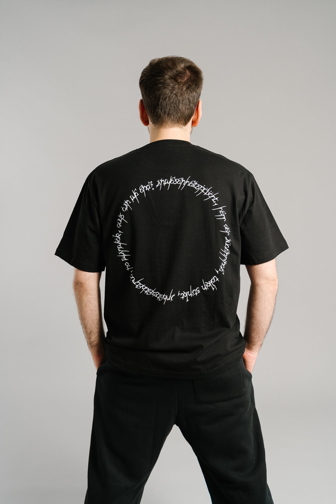 Schwarzes T-Shirt mit großem weißen Rückendruck in kreisförmiger Anordnung. Der Print besteht aus handschriftlich anmutenden Buchstaben, die einen geschlossenen Kreis auf der Rückenmitte bilden. Das Shirt hat einen lockeren Schnitt und besteht aus weichem Baumwollstoff – ein modernes Streetwear-Design mit grafischem Akzent.