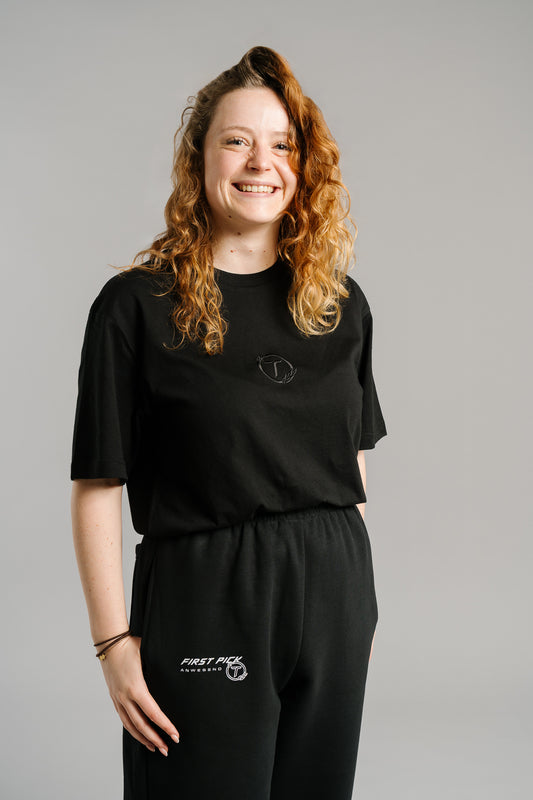 Schwarzes T-Shirt mit dezentem, schwarzem Sticklogo auf der Brust. Das Logo zeigt ein stilisiertes „T“ in einem Lorbeerkranz. Das Shirt besteht aus weicher Baumwolle, hat einen geraden, leicht lockeren Schnitt und verkörpert einen minimalistischen Streetwear-Stil.