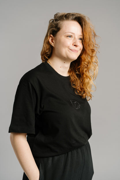 Schwarzes T-Shirt mit feinem Ton-in-Ton-Sticklogo auf der Brust. Das gestickte Emblem zeigt ein stilisiertes „T“ in einem Kreis mit Lorbeerzweigen. Das Shirt hat einen geraden, leicht lockeren Schnitt und besteht aus glatter Baumwolle – schlichtes, hochwertiges Design mit sportivem Charakter.