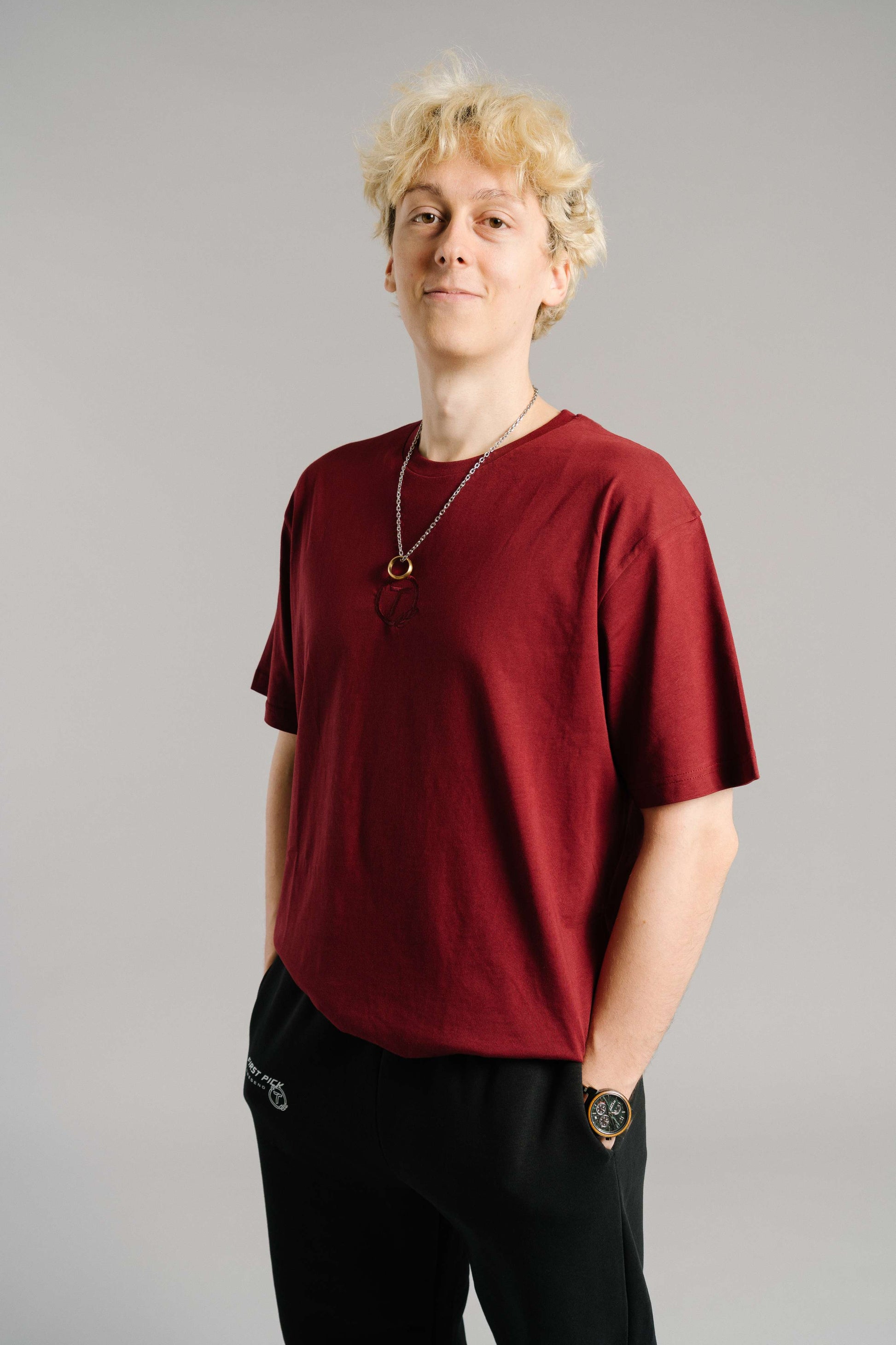 Ein burgunderrotes T-Shirt aus weicher Baumwolle mit tonaler Stickerei des „T“-Logos im Lorbeerkranz auf der Brust. Das Shirt hat einen lockeren, geraden Schnitt mit kurzen Ärmeln und einem Rundhalsausschnitt. Es wird lässig getragen und vermittelt einen modernen, minimalistischen Streetwear-Look.