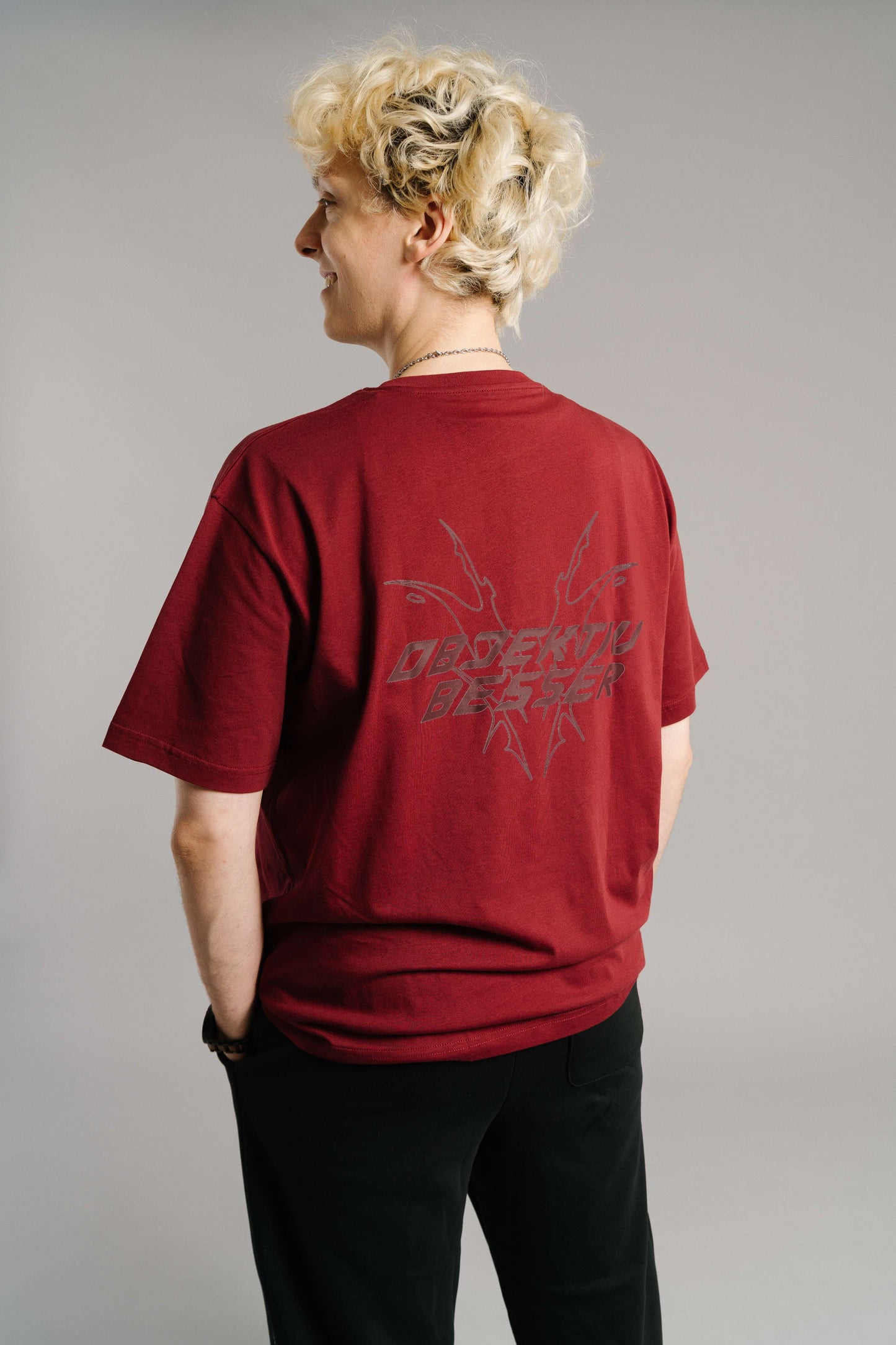 Rückansicht eines burgunderroten T-Shirts mit großem, dunkelgrauem Rückendruck. Das Motiv zeigt den Schriftzug „OBJEKTIV BESSER“ in kantiger, grafischer Typografie, eingerahmt von abstrakten, spitzen Linien, die an Flammen oder Flügel erinnern. Das Shirt hat einen lockeren Schnitt aus weichem Baumwollstoff und kurze Ärmel.
