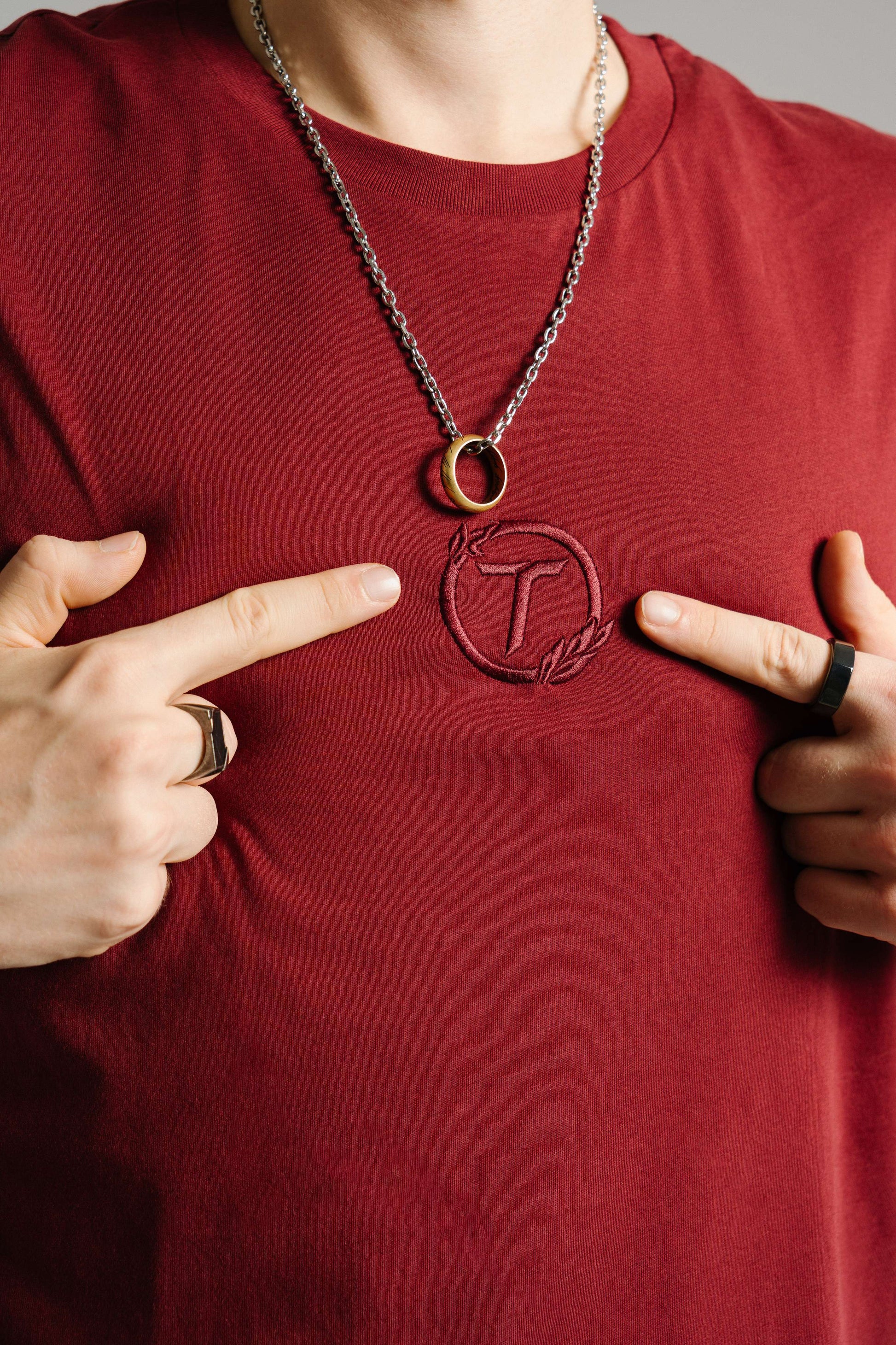 Nahaufnahme eines burgunderroten T-Shirts aus Baumwolle mit tonaler Stickerei des „T“-Logos im Lorbeerkranz auf der Brust. Zwei Finger zeigen auf das gestickte Emblem, das mittig unterhalb einer silbernen Halskette mit goldenem Ringanhänger liegt. Der Fokus liegt auf der Logo-Stickerei und der feinen Stoffstruktur.