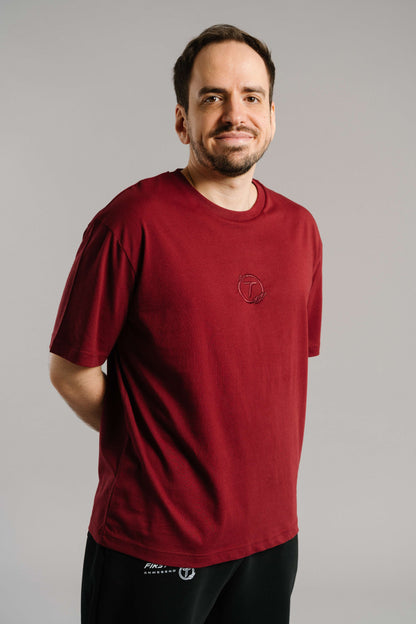 Ein burgunderrotes T-Shirt mit tonaler Stickerei des „T“-Logos im Lorbeerkranz auf der Brust. Das Shirt hat einen geraden Schnitt, kurze Ärmel und besteht aus weicher Baumwolle. Es wird locker getragen und vermittelt einen cleanen, minimalistischen Look.