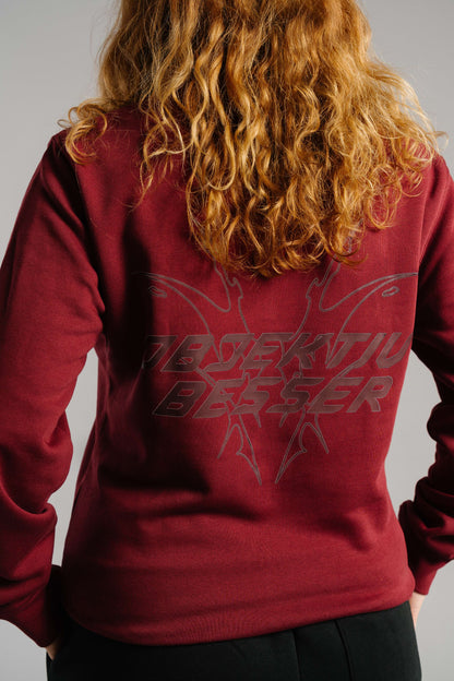 Ein burgunderrotes Sweatshirt mit großem Rückendruck. Der Druck zeigt den Schriftzug „OBJEKTIV BESSER“ in grauer, kantiger Typografie, kombiniert mit einer grafischen Linienillustration im Tribal-Stil. Der Stoff wirkt weich und dicht. Das Bild zeigt den oberen Rücken und einen Teil der Arme einer Person mit lockigen, kupferblonden Haaren, die den Print teilweise überdecken.