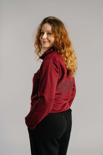 Ein burgunderrotes Sweatshirt mit kurzem Reißverschluss und lockerem Schnitt. Auf der Rückseite befindet sich ein großflächiger, dunkler Schriftzug mit grafischem Motiv. Der Stoff wirkt weich und gemütlich. Die Person steht leicht seitlich zur Kamera, blickt mit einem dezenten Lächeln über die Schulter und trägt das Sweatshirt lässig zu einer schwarzen Hose in neutraler Studioumgebung.