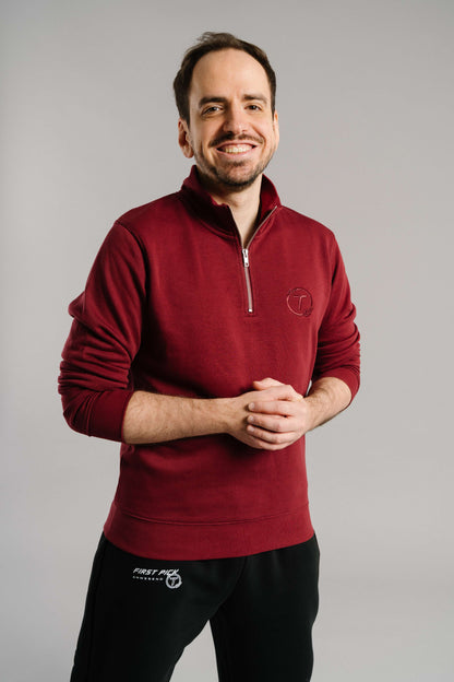 Ein burgunderroter Sweatshirt-Pullover mit kurzem Reißverschluss am Kragen. Auf der linken Brust befindet sich eine ton-in-ton Stickerei mit dem Buchstaben „T“ in einem Lorbeerkranz. Der Stoff wirkt weich und leicht strukturiert. Die Person trägt den Pullover leger, mit leicht angewinkelten Armen und einem freundlichen Lächeln in neutraler Studioumgebung.