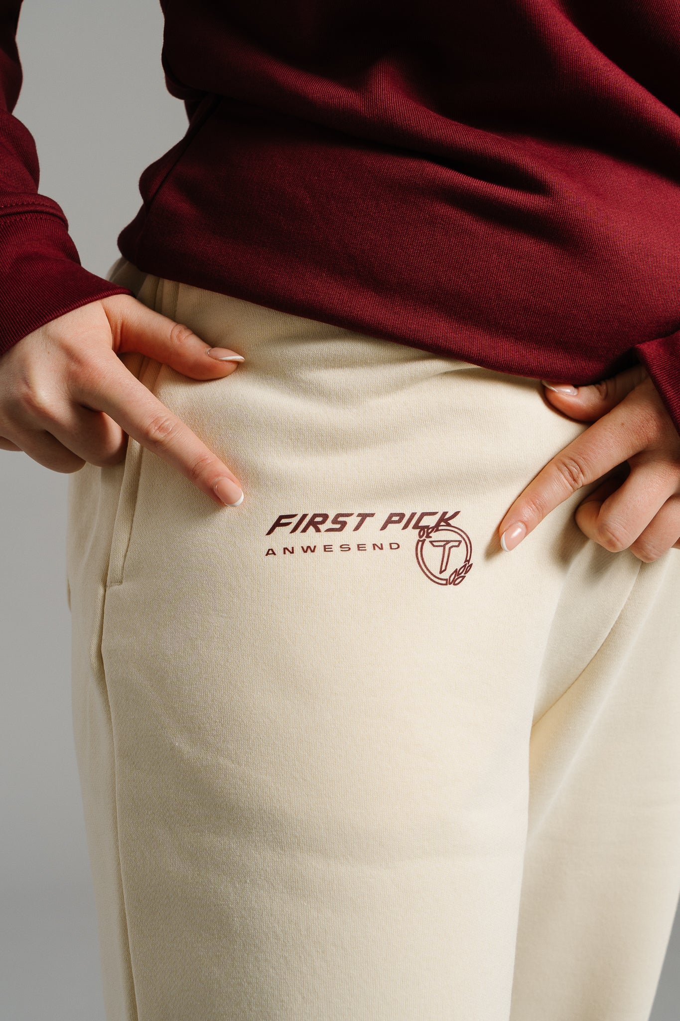 Detailaufnahme einer beigefarbenen Jogginghose aus weichem Sweatstoff. Auf der linken Vorderseite ist ein braunroter Print mit der Aufschrift „FIRST PICK ANWESEND“ und einem runden Logo mit stilisiertem Buchstaben „T“ zu sehen. Zwei Finger zeigen auf den Aufdruck, der sich oberhalb der Hosentasche befindet.