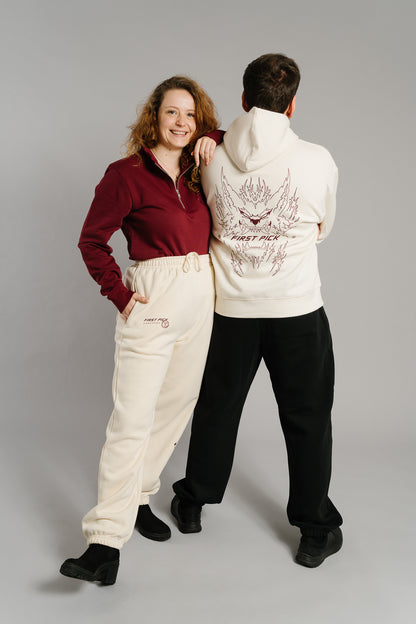 Beigefarbene Jogginghose mit lockerer Passform und elastischen Bündchen an Bund und Knöcheln. Auf der linken Vorderseite befindet sich ein braunroter Aufdruck mit der Aufschrift „FIRST PICK ANWESEND“ und einem runden Logo. Die Hose besteht aus weichem Sweatstoff und hat seitliche Eingrifftaschen.