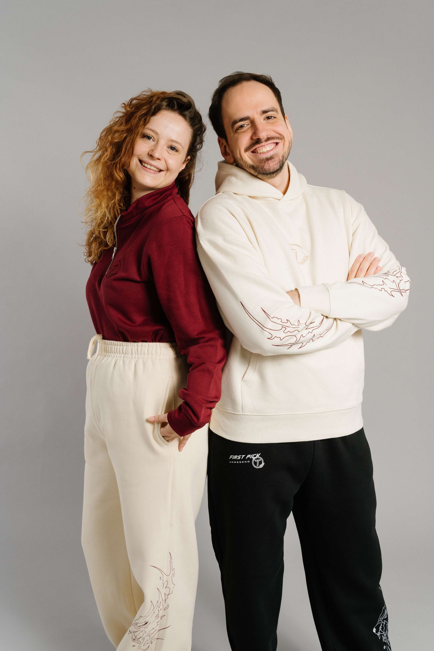 Ein burgunderrotes Sweatshirt mit kurzem Reißverschluss und gesticktem Brustlogo. Das Sweatshirt hat eine weiche, strukturierte Oberfläche und sitzt locker. Die Person trägt es kombiniert mit einer hellen Jogginghose und steht seitlich mit den Händen in den Hosentaschen. Neben ihr steht eine weitere Person in einem cremefarbenen Hoodie; beide lächeln und lehnen sich Rücken an Rücken, wodurch das Sweatshirt im Vordergrund betont wird.