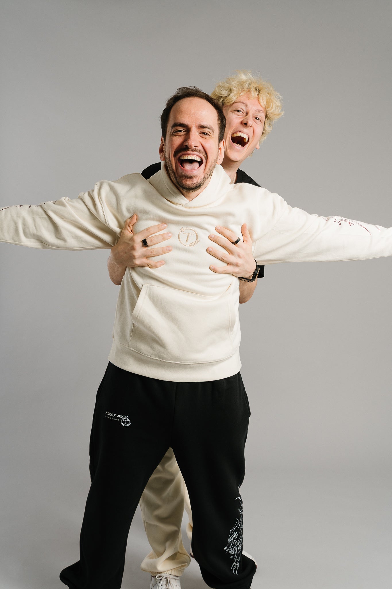 Zwei lachende Personen posieren humorvoll voreinander. Die vordere trägt einen beigefarbenen Hoodie mit kleinem Logo auf der Brust und flammenförmigen Linienmotiven auf den Ärmeln. Der Hoodie hat eine weiche, lockere Passform mit Kängurutasche und betont den gemütlichen Streetwear-Stil. Die hintere Person greift ihr spielerisch an die Brust und lacht laut, wodurch die Szene lebendig und witzig wirkt.