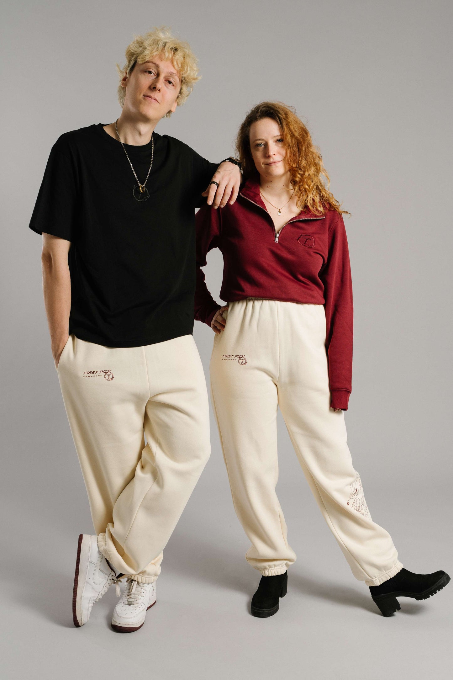 Ein burgunderrotes Sweatshirt mit kurzem Reißverschluss und tonaler Bruststickerei des „T“-Logos im Lorbeerkranz. Das Sweatshirt hat eine weiche Struktur und wird locker in eine cremefarbene Jogginghose mit elastischem Bund und seitlichen Taschen getragen. Die Person steht neben jemandem im schwarzen T-Shirt und trägt das Sweatshirt mit leicht geöffnetem Kragen. Der Fokus liegt auf der sportlich-lässigen Kombination des burgunderfarbenen Oberteils.