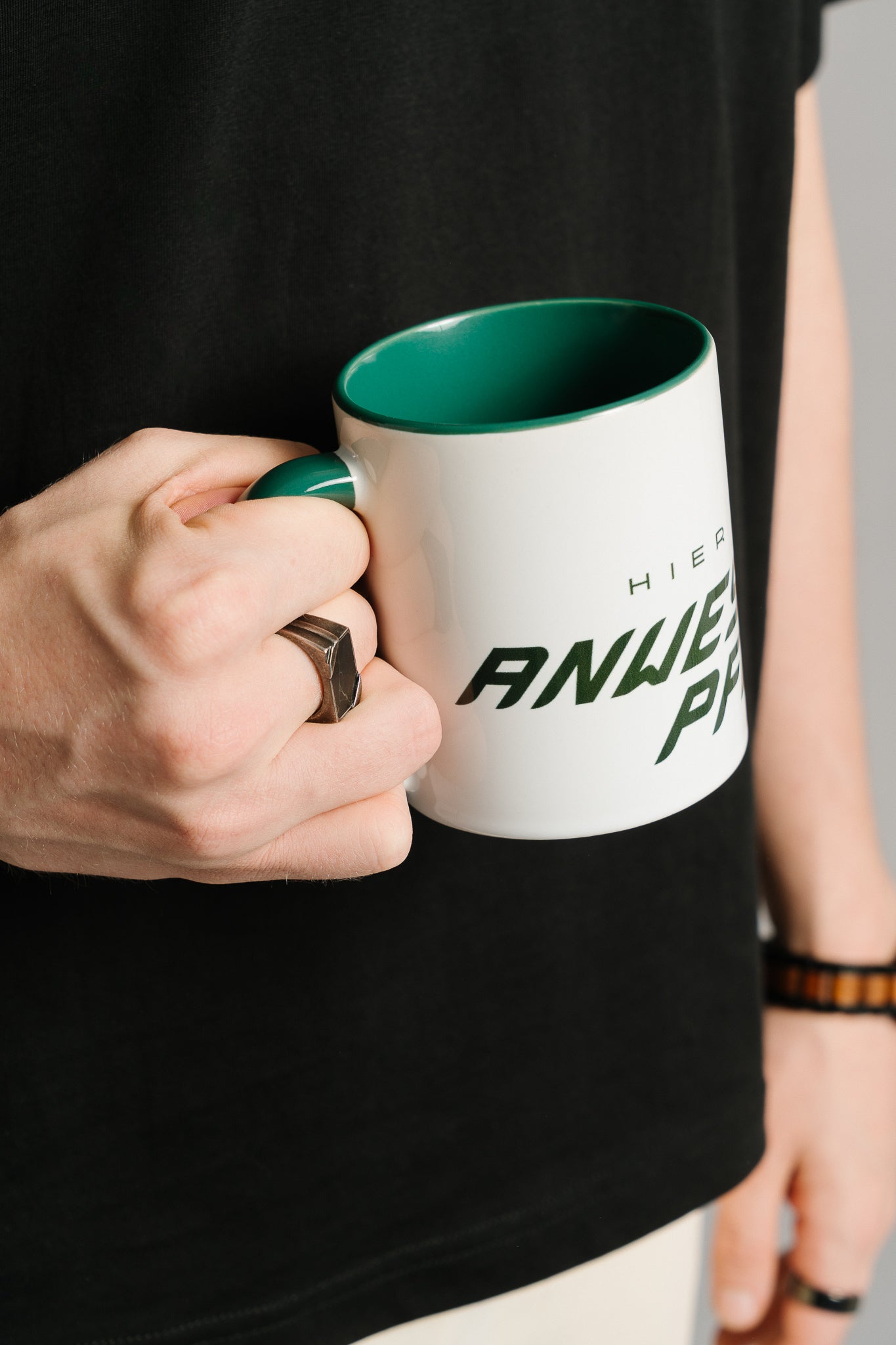 Nahaufnahme einer Hand, die eine weiße Keramiktasse mit grünem Innenbereich und Henkel hält. Auf der Tasse ist ein dunkelgrüner Schriftzug zu sehen, der teilweise den Text „HIER HERRSCHT ANWESENHEITSPFLICHT“ zeigt. Die Person trägt ein schwarzes T-Shirt sowie Ringe und ein Armband; der Hintergrund ist neutral gehalten.