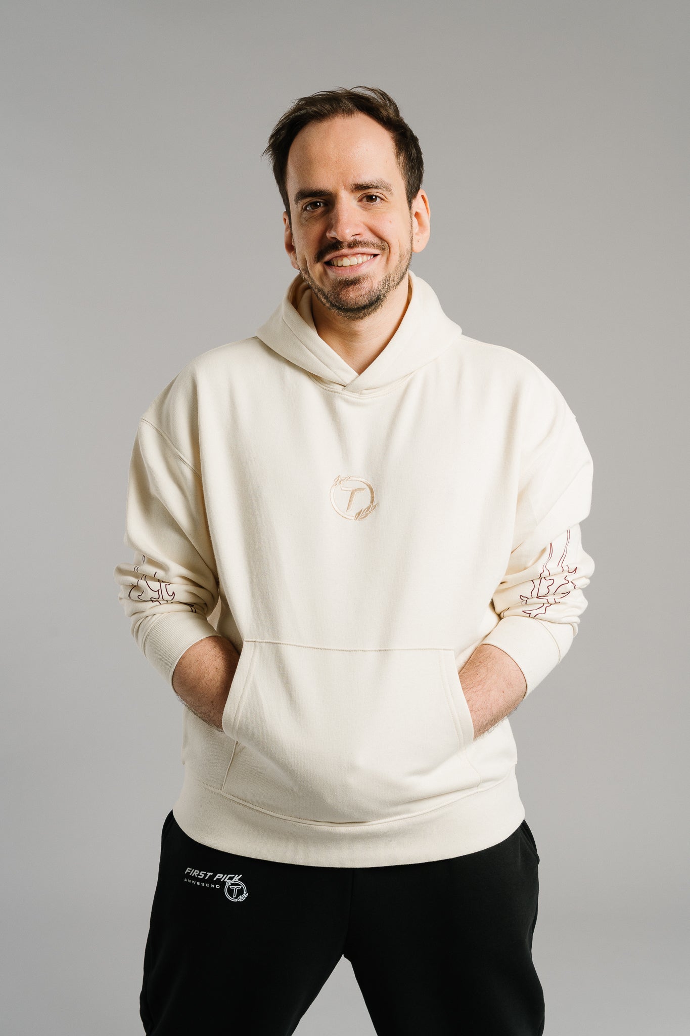 Eine lächelnde Person steht frontal in neutraler Pose vor hellem Hintergrund. Sie trägt einen beigefarbenen Hoodie mit kleinem, rundem Logo auf der Brust und dezenten, flammenförmigen Linienmotiven auf den Ärmeln. Der Hoodie hat eine Kängurutasche und eine lockere, bequeme Passform, die den modernen Streetwear-Stil betont. Darunter ist eine schwarze Jogginghose mit weißem Schriftzug zu sehen.