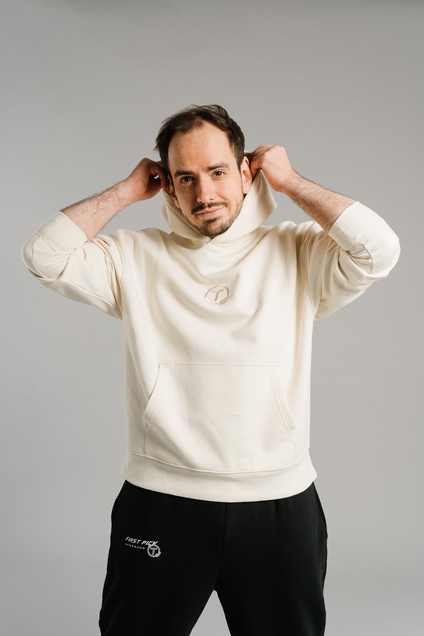 Eine Person steht vor hellem Hintergrund und greift mit beiden Händen an die Kapuze eines beigefarbenen Hoodies. Der Hoodie besteht aus dickem, weichem Stoff und zeigt auf der Brust ein rundes Logo in passendem Beigeton. Die weite Passform mit Kängurutasche und breiten Bündchen verleiht dem Kleidungsstück einen modernen, gemütlichen Look, der für lässige Streetwear steht.