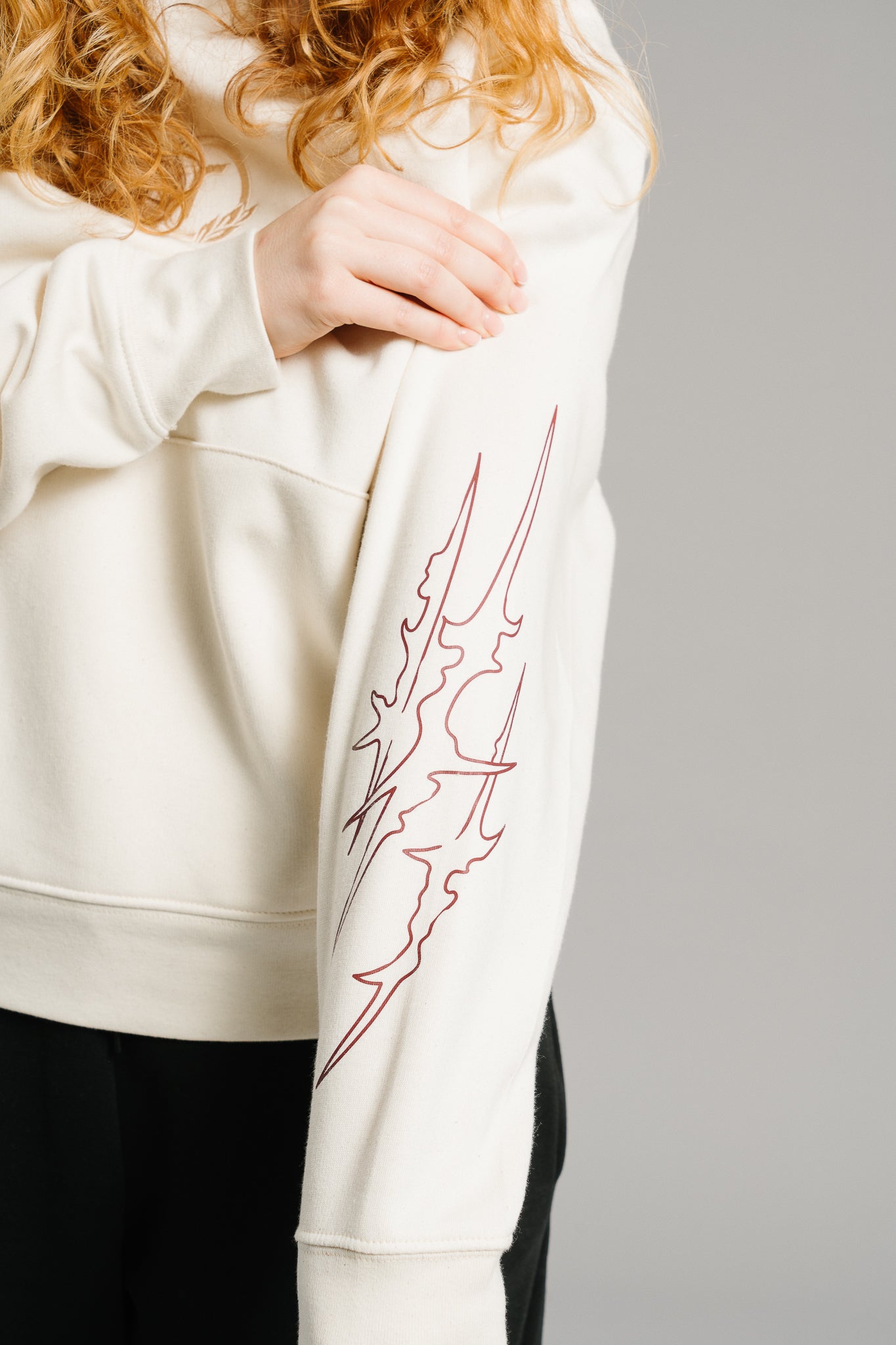 Detailaufnahme eines beigefarbenen Hoodies. Auf dem Ärmel ist ein filigranes, rotbraunes Linienmotiv in Form abstrakter Flammen zu sehen. Der weiche Stoff und die saubere Verarbeitung betonen die hochwertige Qualität des Hoodies, während das markante Ärmel-Design dem schlichten Look eine dynamische Note verleiht.