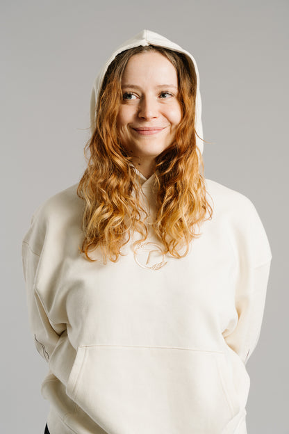 Eine Person trägt einen beigefarbenen Hoodie mit aufgesetzter Kapuze und lächelt leicht in neutralem Studiolicht. Auf der Brust ist ein rundes Logo mit stilisiertem Buchstaben „T“ ton-in-ton gestickt. Der weiche Stoff und die lockere Passform unterstreichen den gemütlichen, modernen Look des Hoodies.