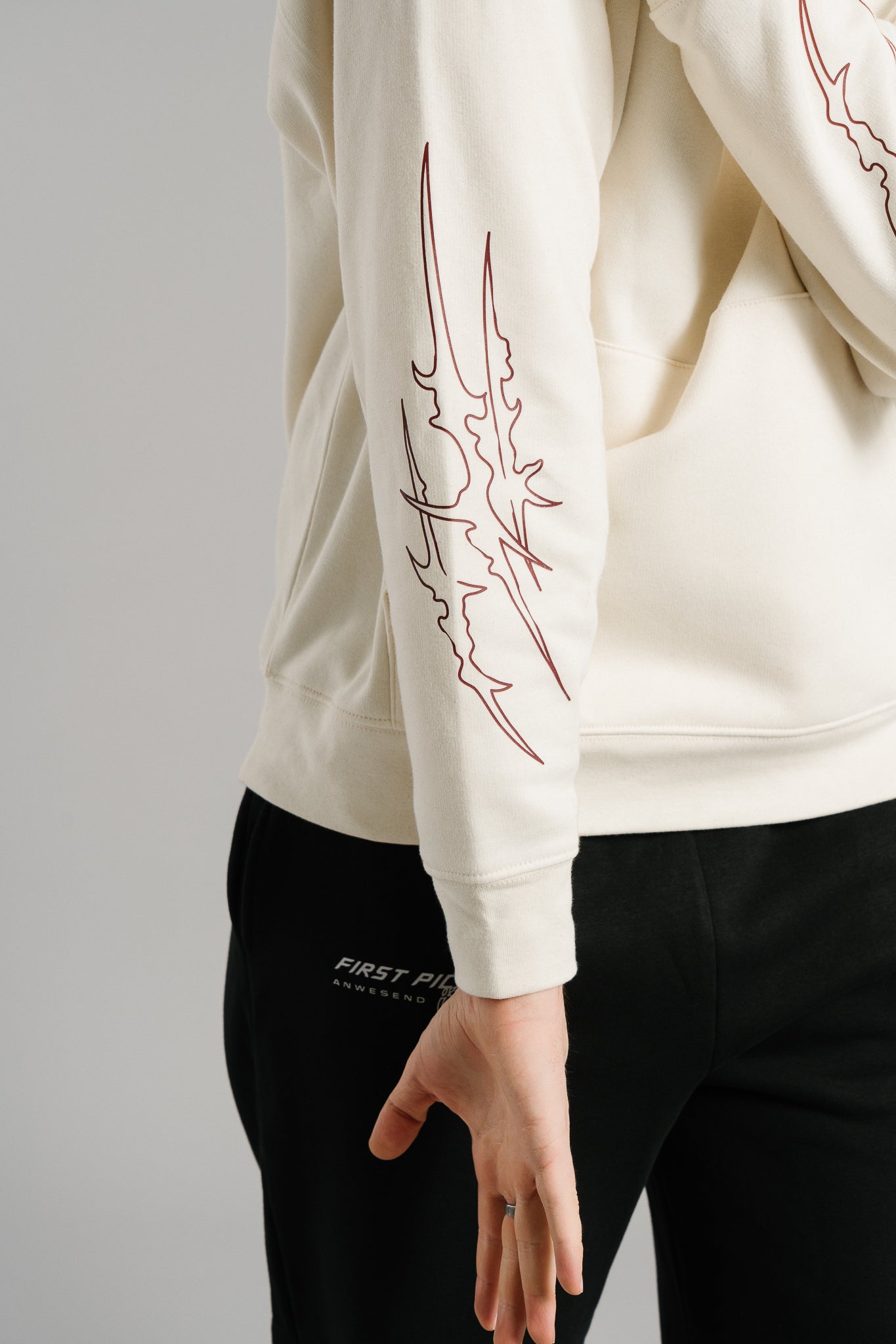 Detailaufnahme eines beigefarbenen Hoodies aus Sweatstoff. Auf dem Ärmel ist ein dunkelrotes, flammenförmiges Linienmotiv aufgedruckt. Der Stoff wirkt weich und strukturiert, der Ärmel endet in einem Bündchen. Im unteren Bildausschnitt ist eine schwarze Jogginghose mit weißem „FIRST PICK ANWESEND“-Print zu sehen.