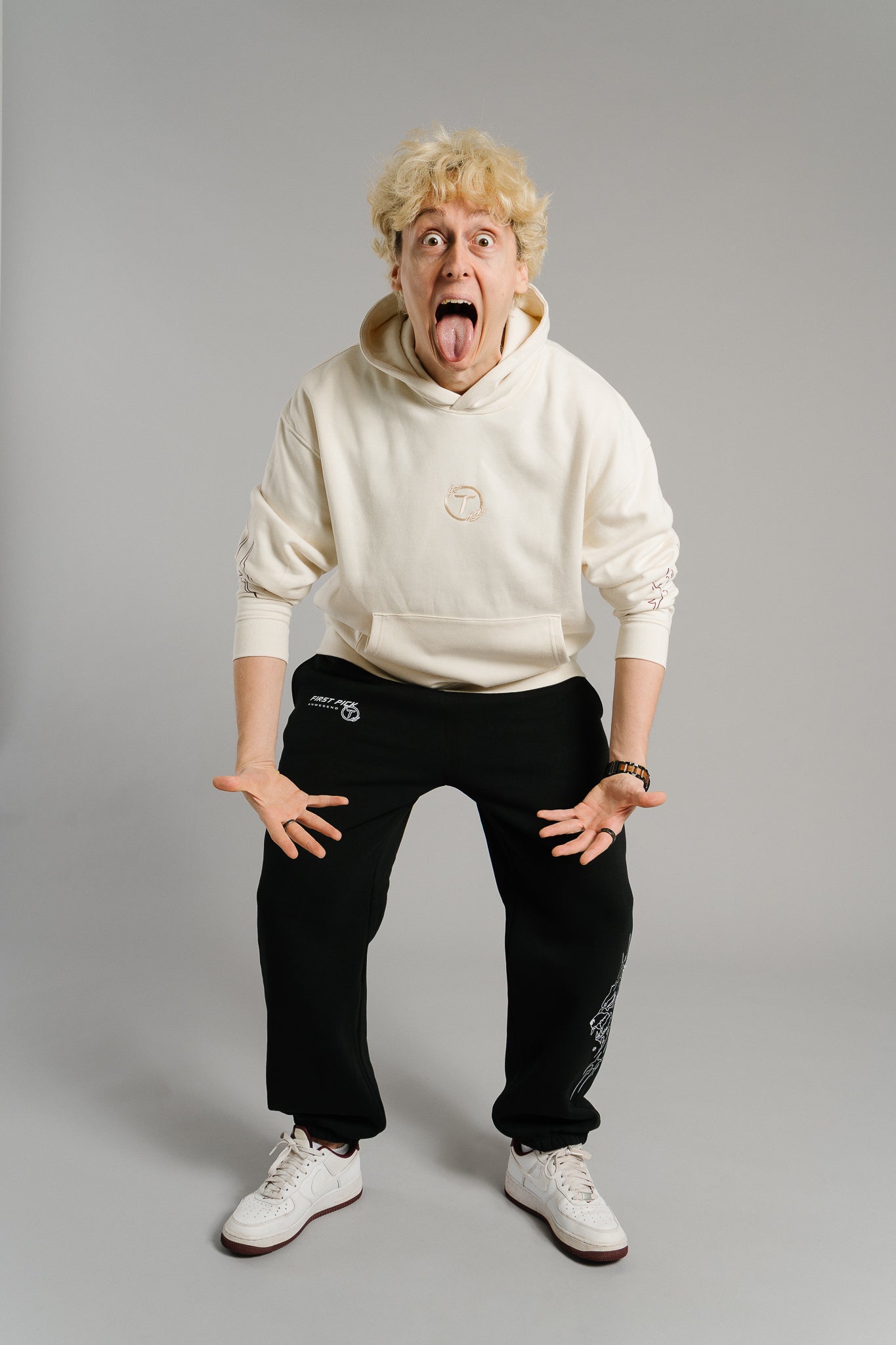 Eine Person steht in humorvoller Pose im Studio und streckt lachend die Zunge heraus. Sie trägt einen beigefarbenen Hoodie mit dezent gesticktem Logo auf der Brust und grafischen Linienmotiven an den Ärmeln. Der Hoodie wirkt weich und hochwertig, während die lockere Passform und die verspielte Haltung den lässigen Streetwear-Charakter unterstreichen.