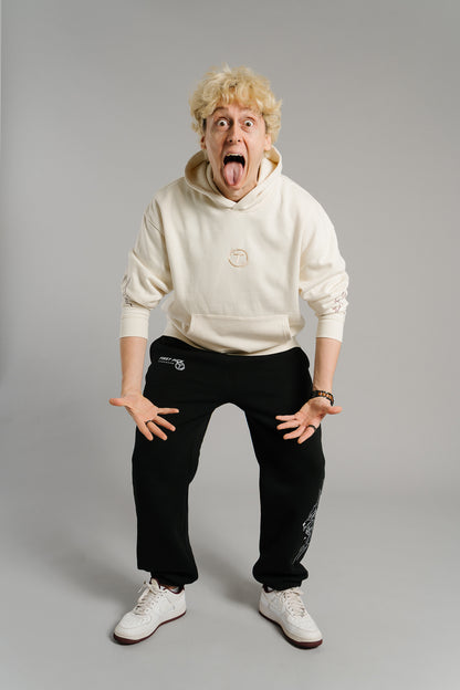 Eine Person steht in humorvoller Pose im Studio und streckt lachend die Zunge heraus. Sie trägt einen beigefarbenen Hoodie mit dezent gesticktem Logo auf der Brust und grafischen Linienmotiven an den Ärmeln. Der Hoodie wirkt weich und hochwertig, während die lockere Passform und die verspielte Haltung den lässigen Streetwear-Charakter unterstreichen.