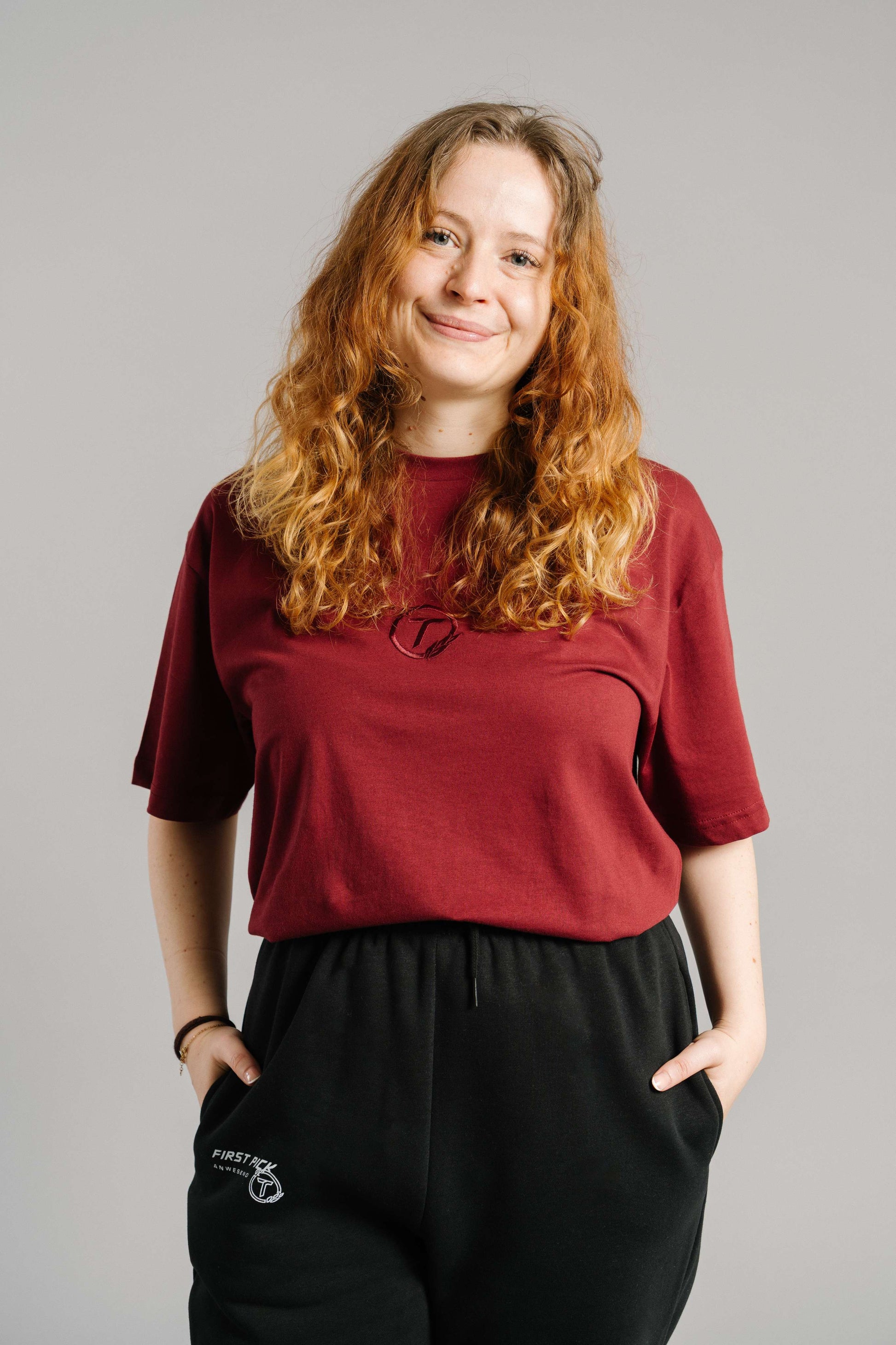 Burgunderrotes T-Shirt mit tonaler Stickerei des „T“-Logos im Lorbeerkranz auf der Brust. Das Shirt hat einen lockeren, geraden Schnitt mit kurzen Ärmeln und Rundhalsausschnitt. Der weiche Baumwollstoff fällt leicht über die Taille und wird zu einer schwarzen Jogginghose kombiniert.